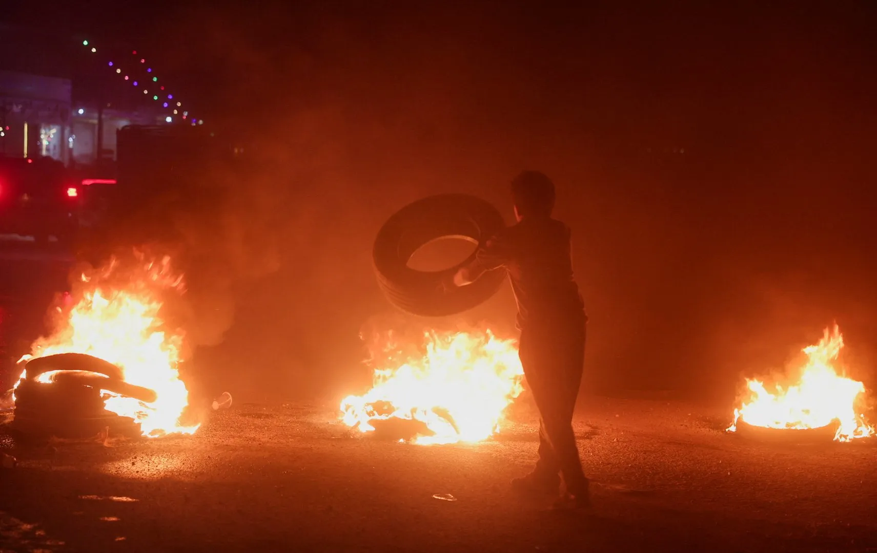 Person carries a tire past burning tires on a dark street at night.