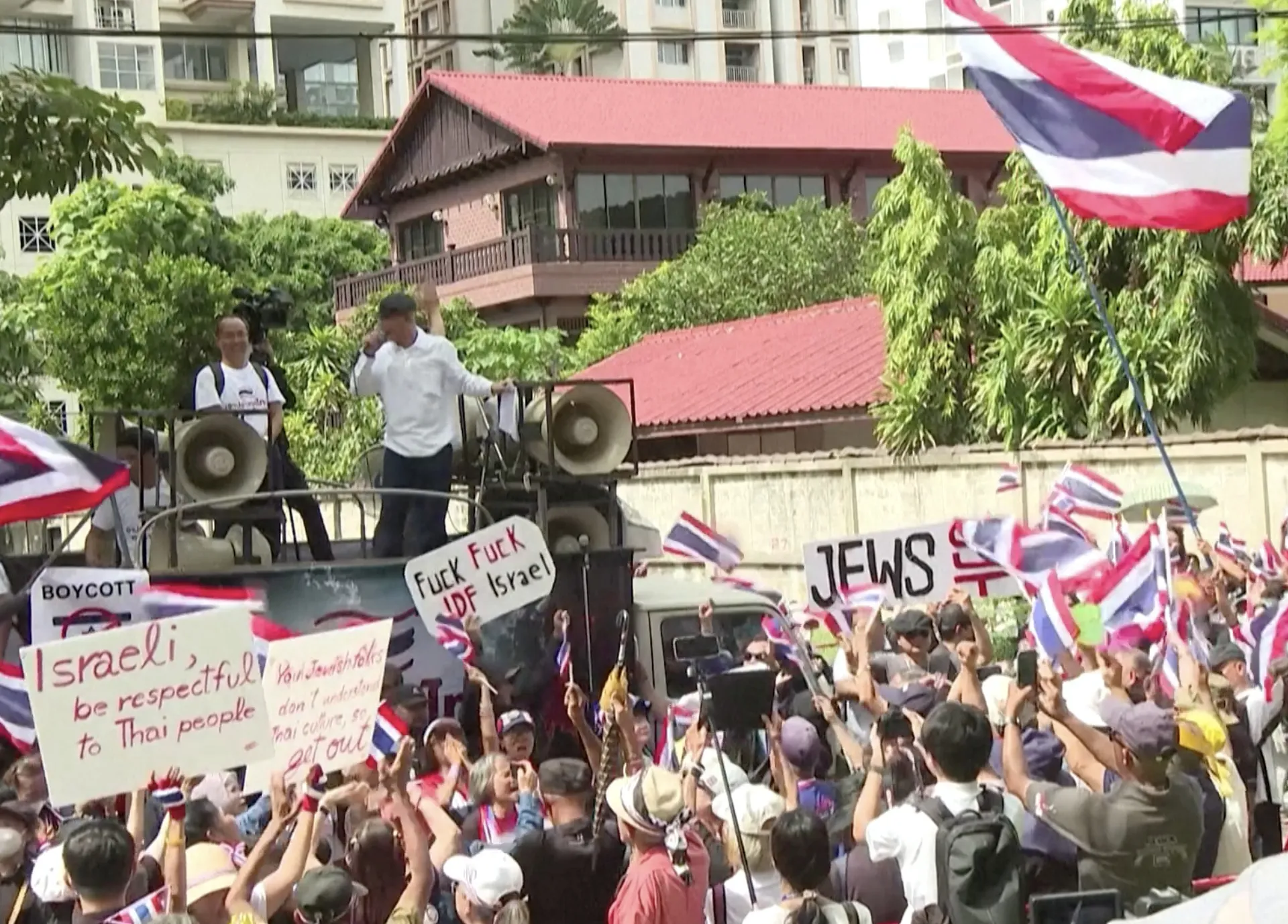 Crowd waving Thai flags gathers before speakers and protest signs outside a building.