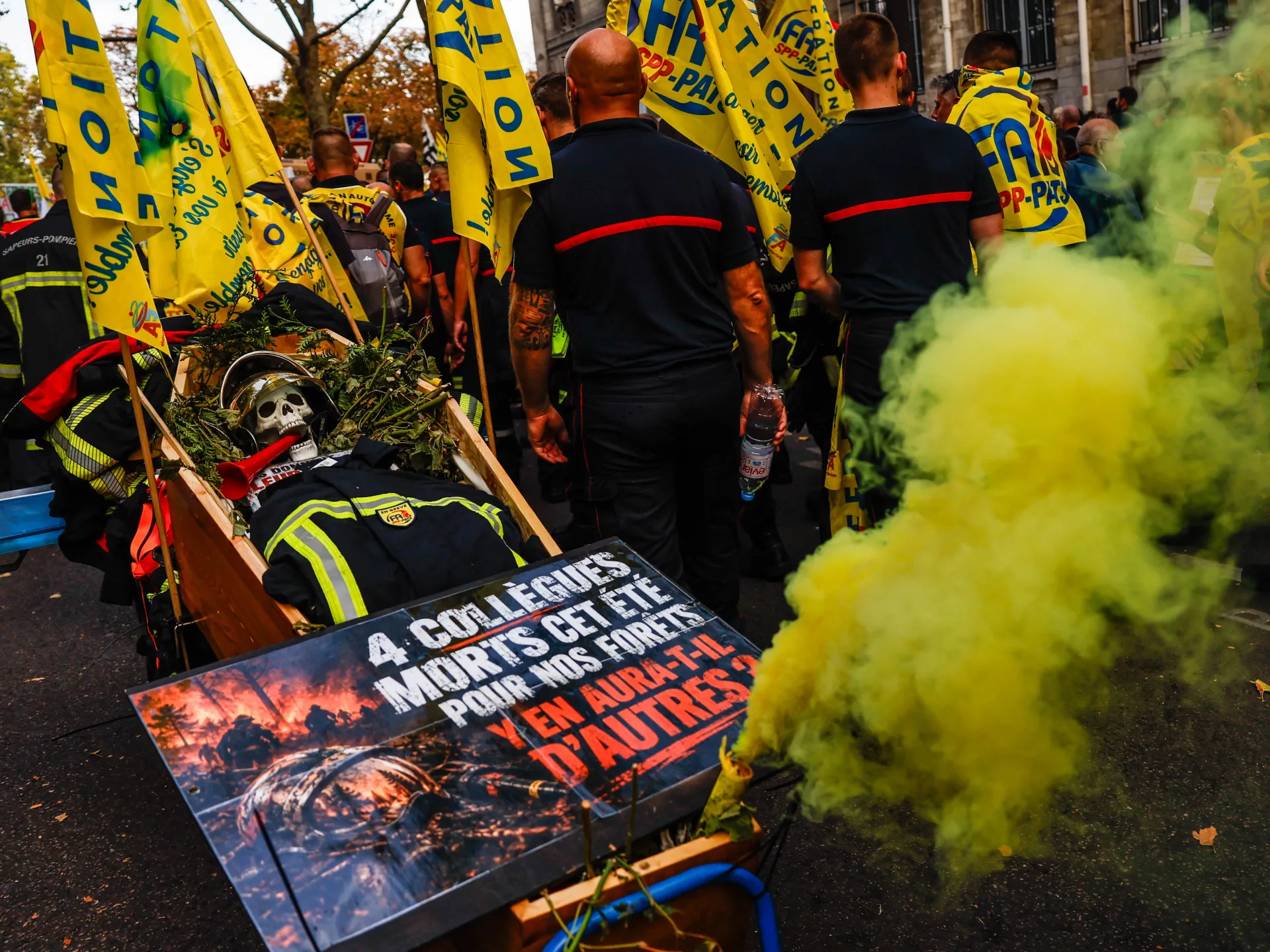 Firefighters march behind yellow union flags past a coffin-shaped display as yellow smoke fills the street.
