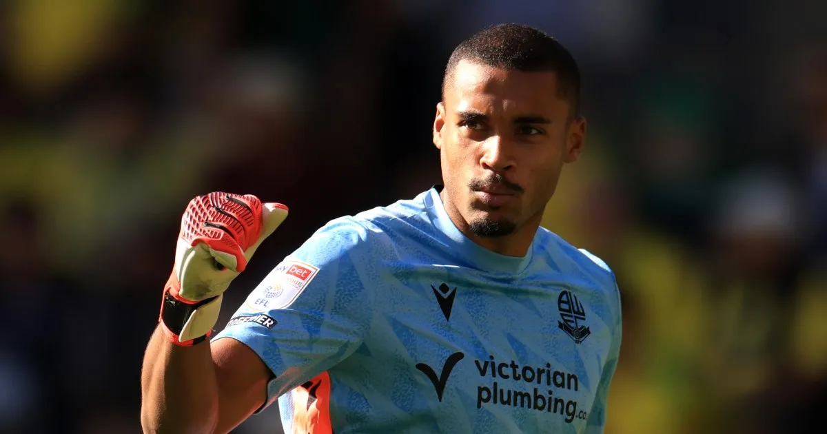 A goalkeeper in a light blue jersey raises a red-and-white gloved hand during a match.