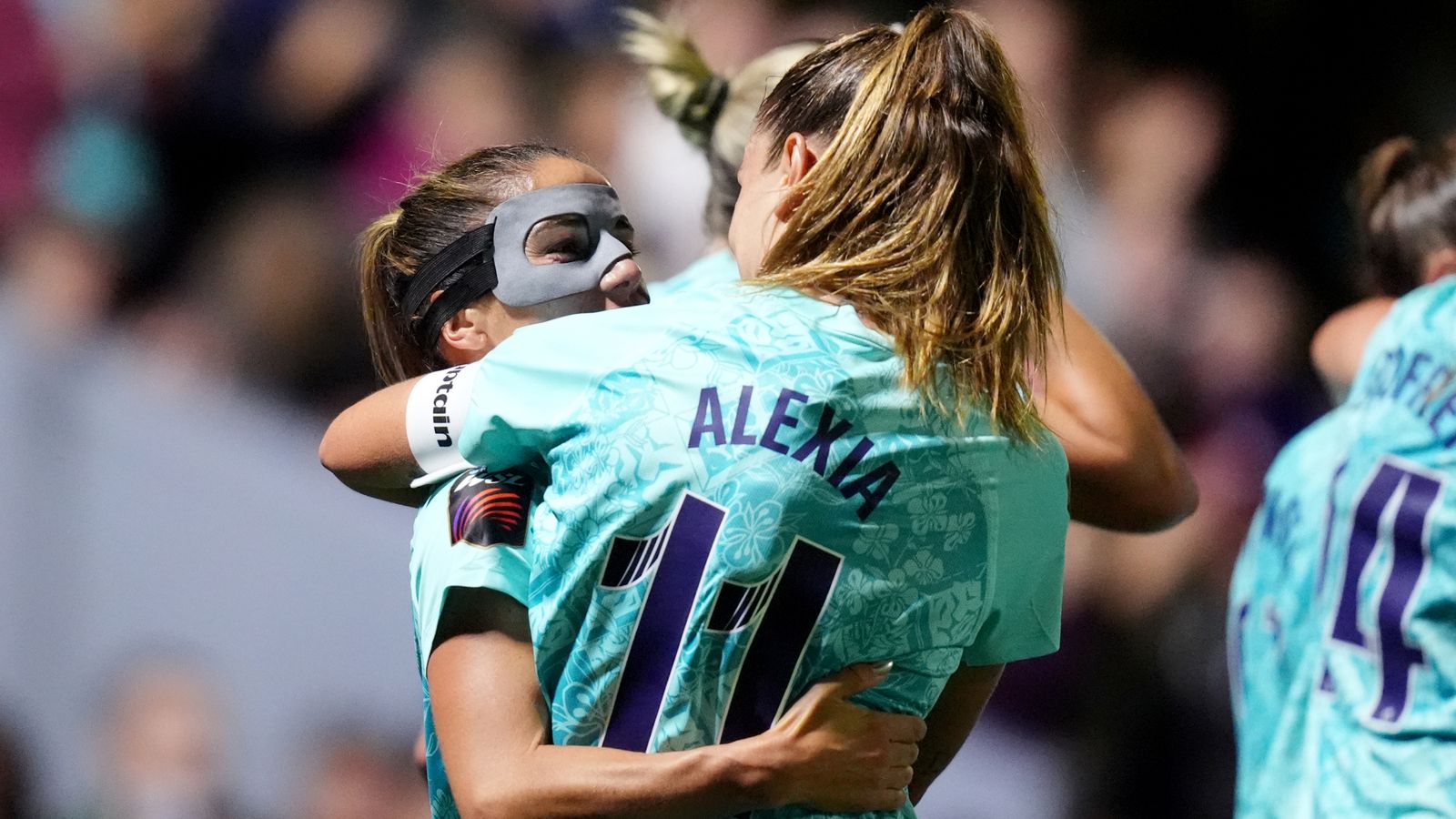 Footballers in turquoise jerseys embrace during a match, with one player wearing a protective face mask.
