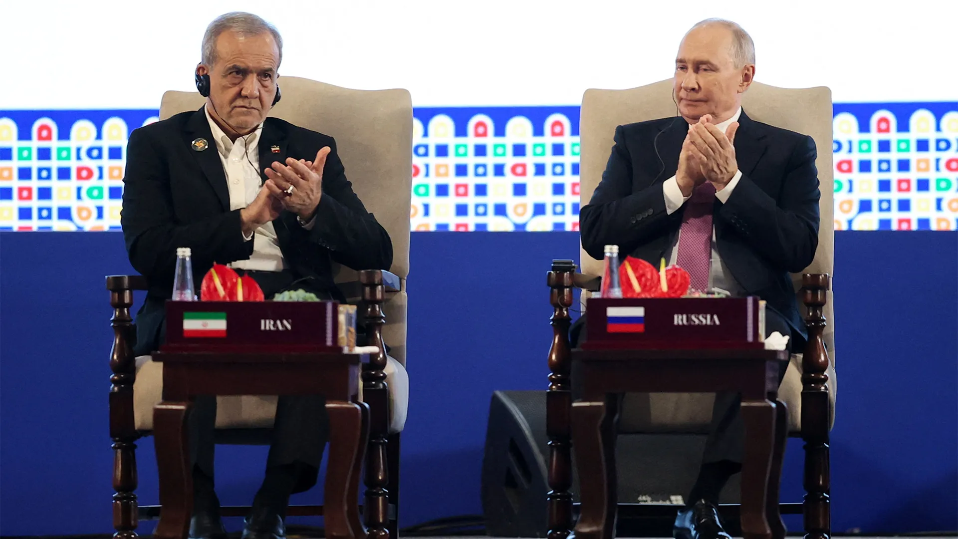 Two suited men seated in armchairs clap beside tables labeled Iran and Russia.