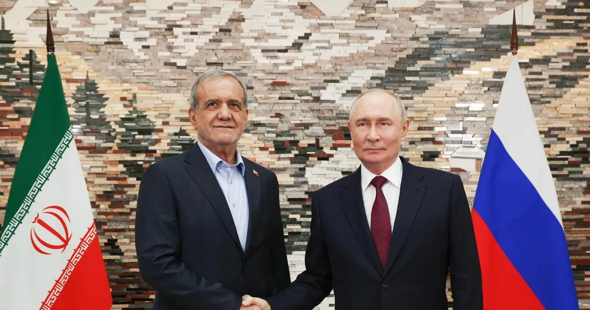 Two suited officials shake hands between Iranian and Russian flags against a stone wall.