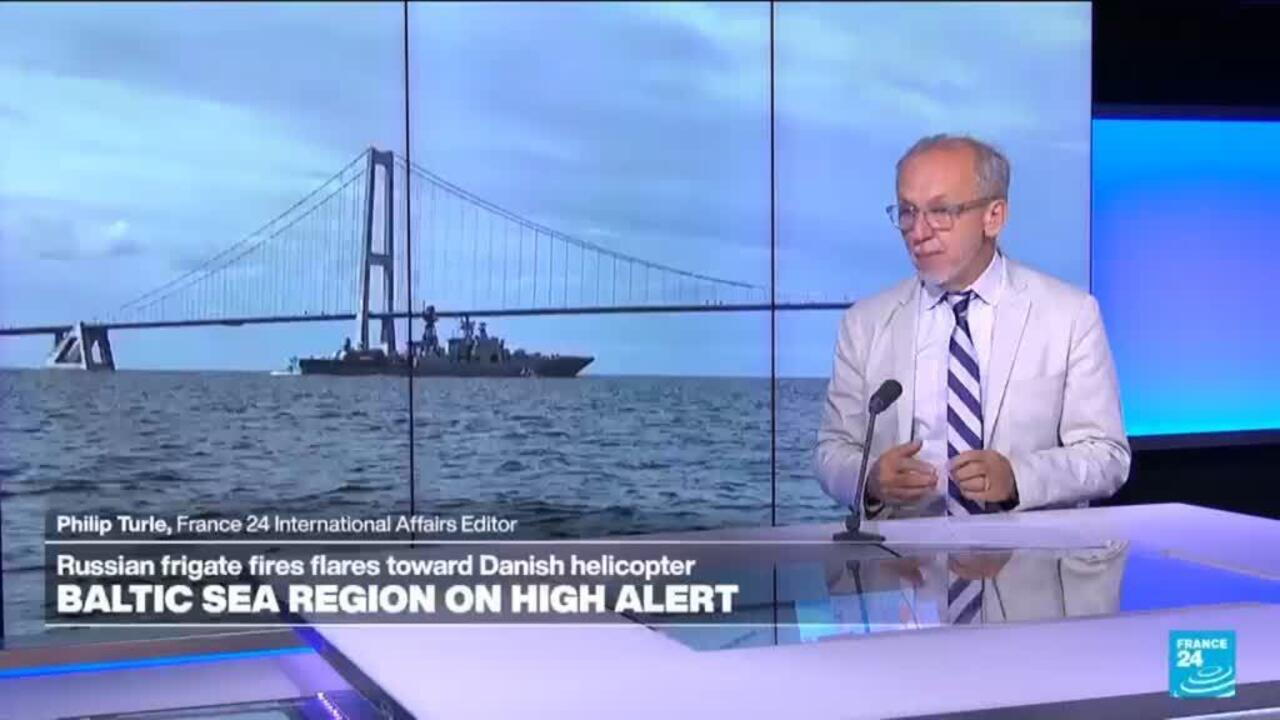Television news presenter beside footage of a warship sailing beneath a suspension bridge