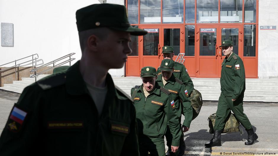 Russian servicemen in green uniforms walk outside a building with red-framed doors.