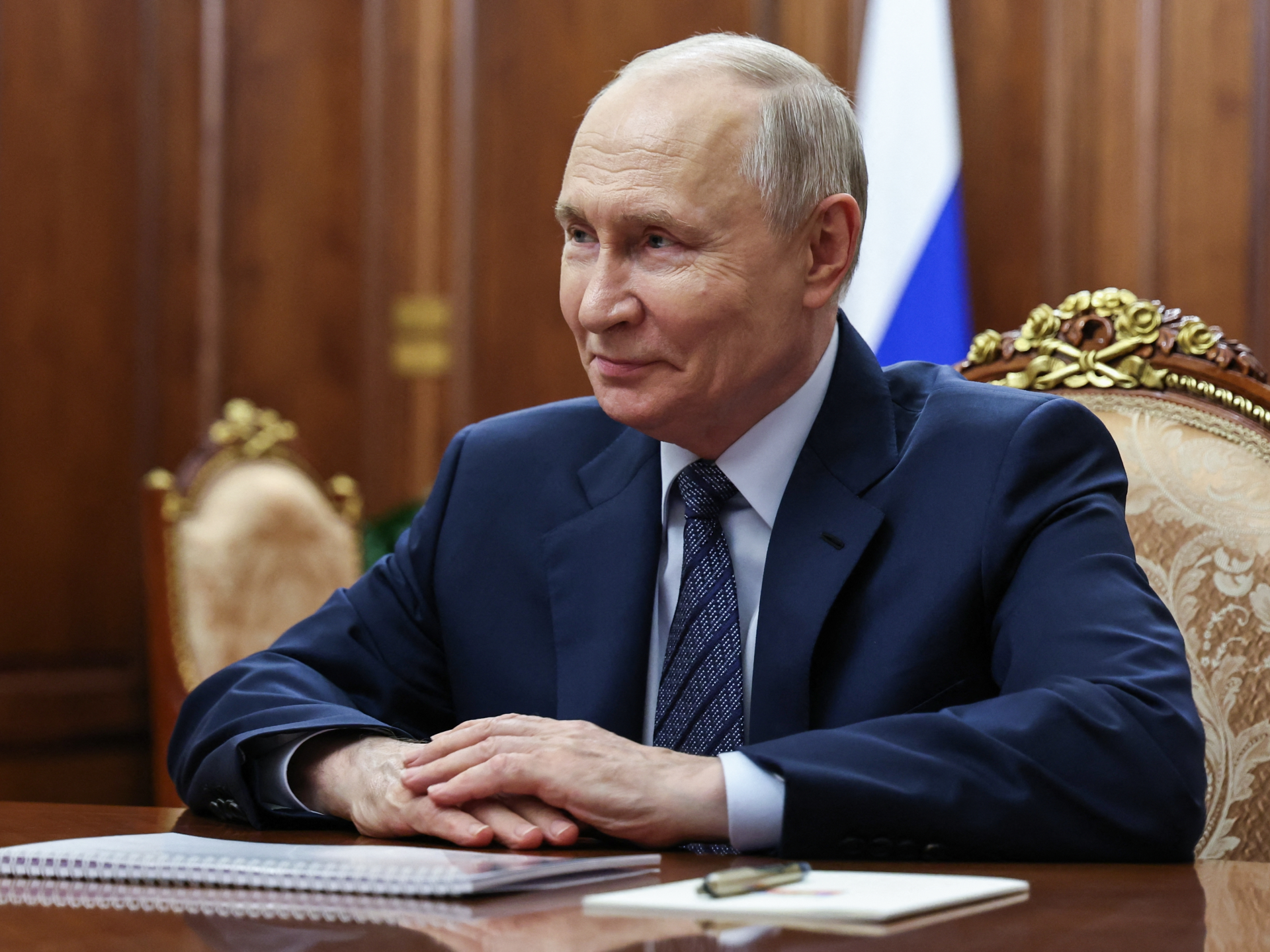 Vladimir Putin sits at a desk in a dark suit inside an ornate room.