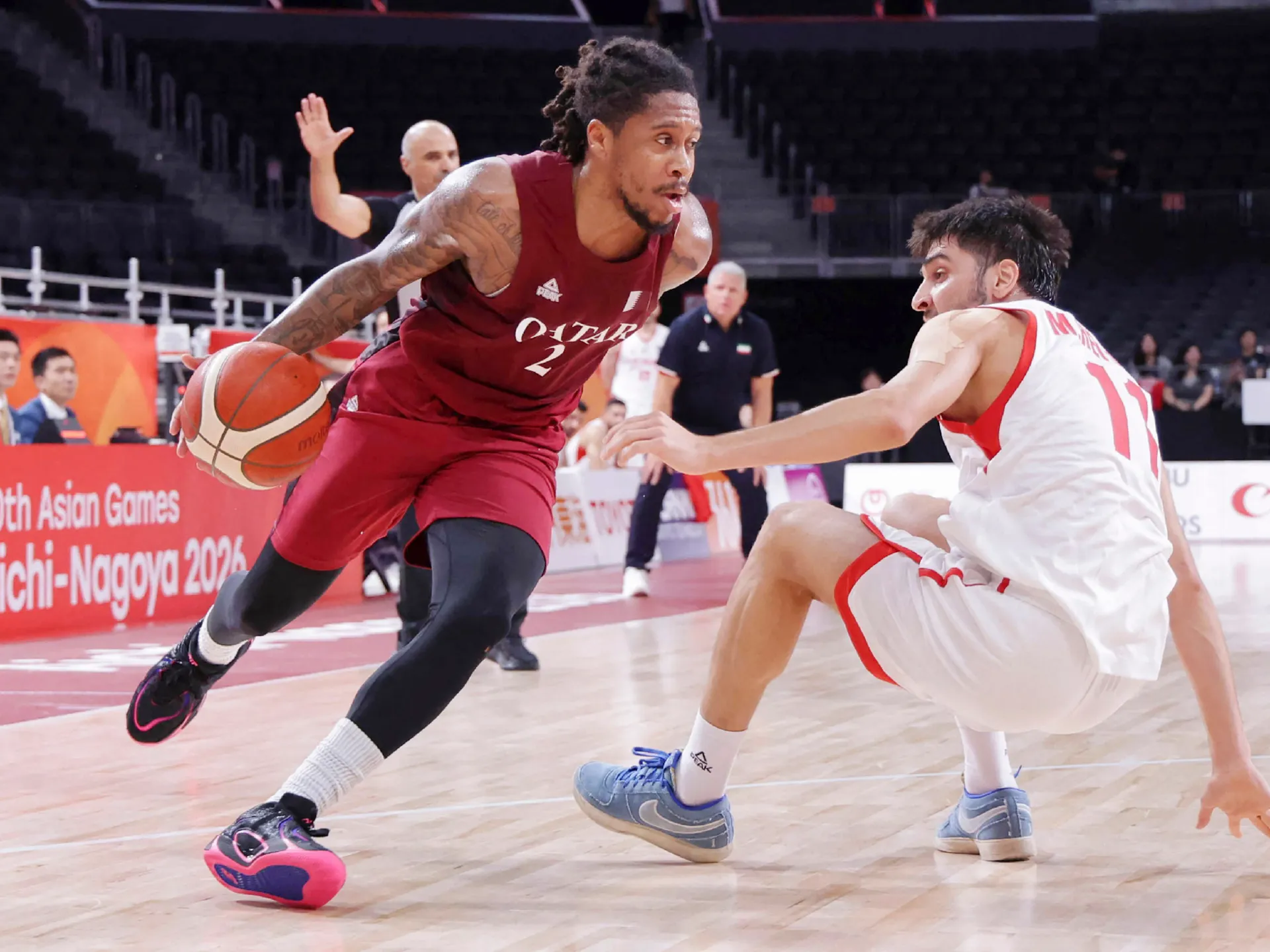 Basketball player in maroon Qatar uniform dribbles past a seated defender in white during a game.