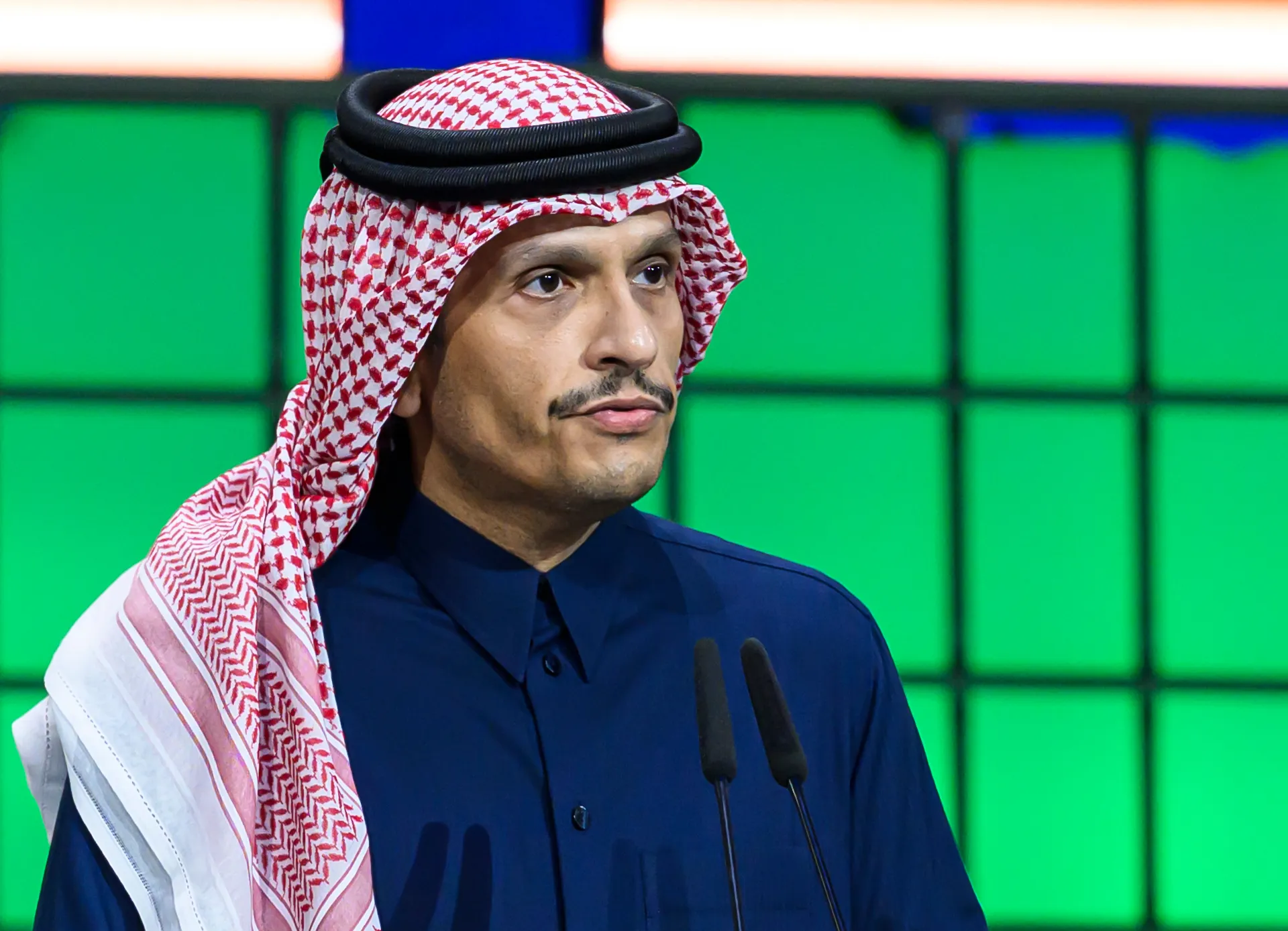 Man in traditional Qatari attire speaking at a podium with two microphones