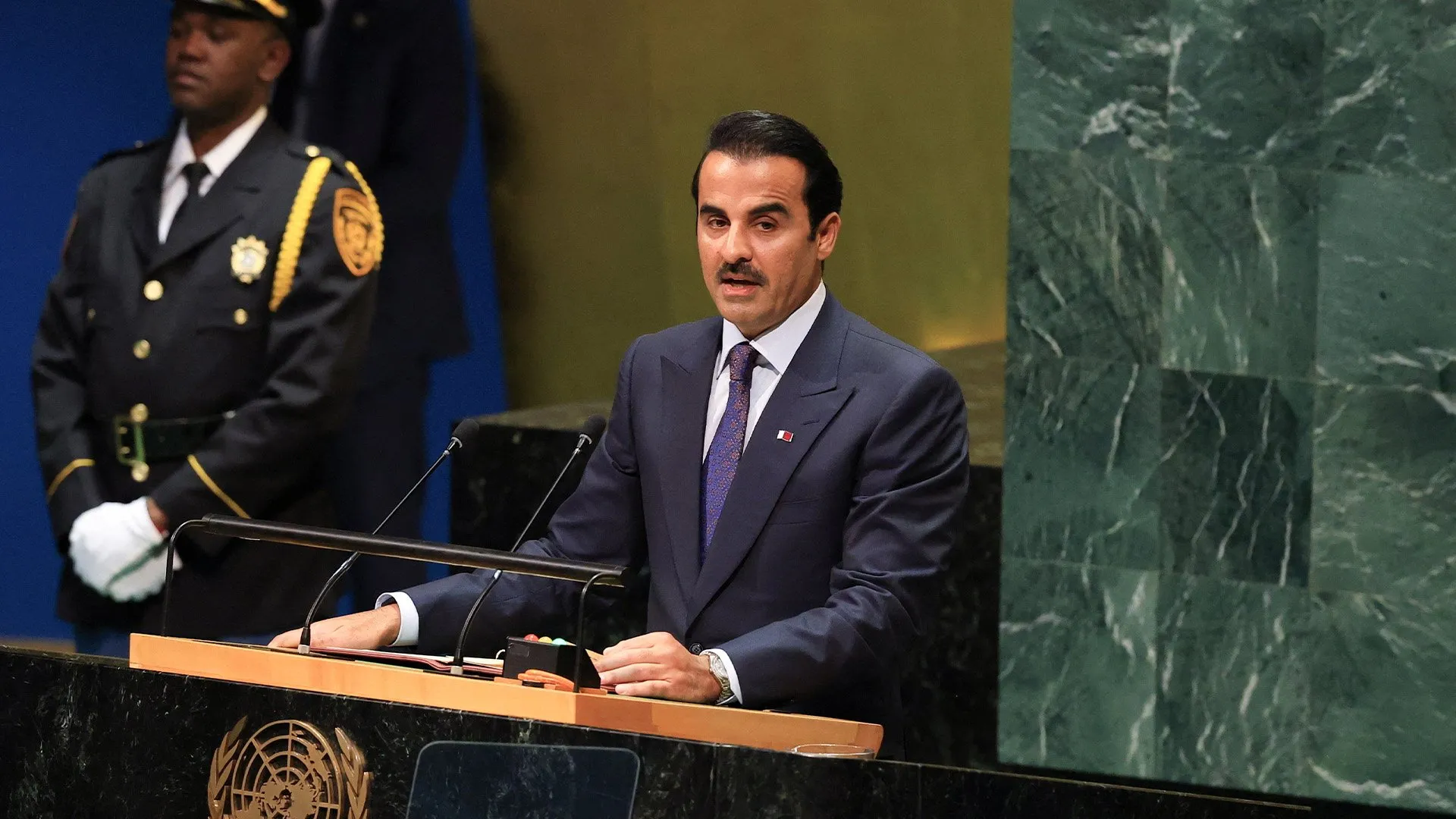 Man in suit speaking at a podium beside a uniformed ceremonial guard.