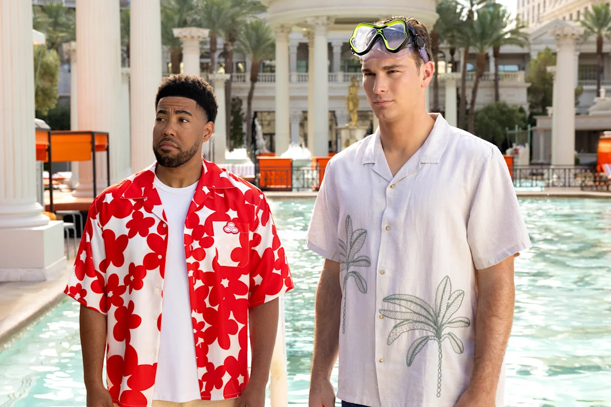 Two men in patterned shirts stand beside a resort pool, one wearing swim goggles.