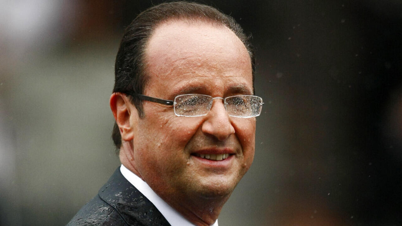 Man wearing glasses and dark suit looks toward camera against a blurred background.