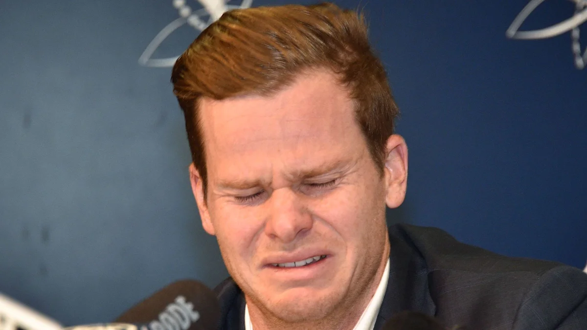 A red-haired man in a dark suit grimaces with his eyes closed as microphones point toward him.