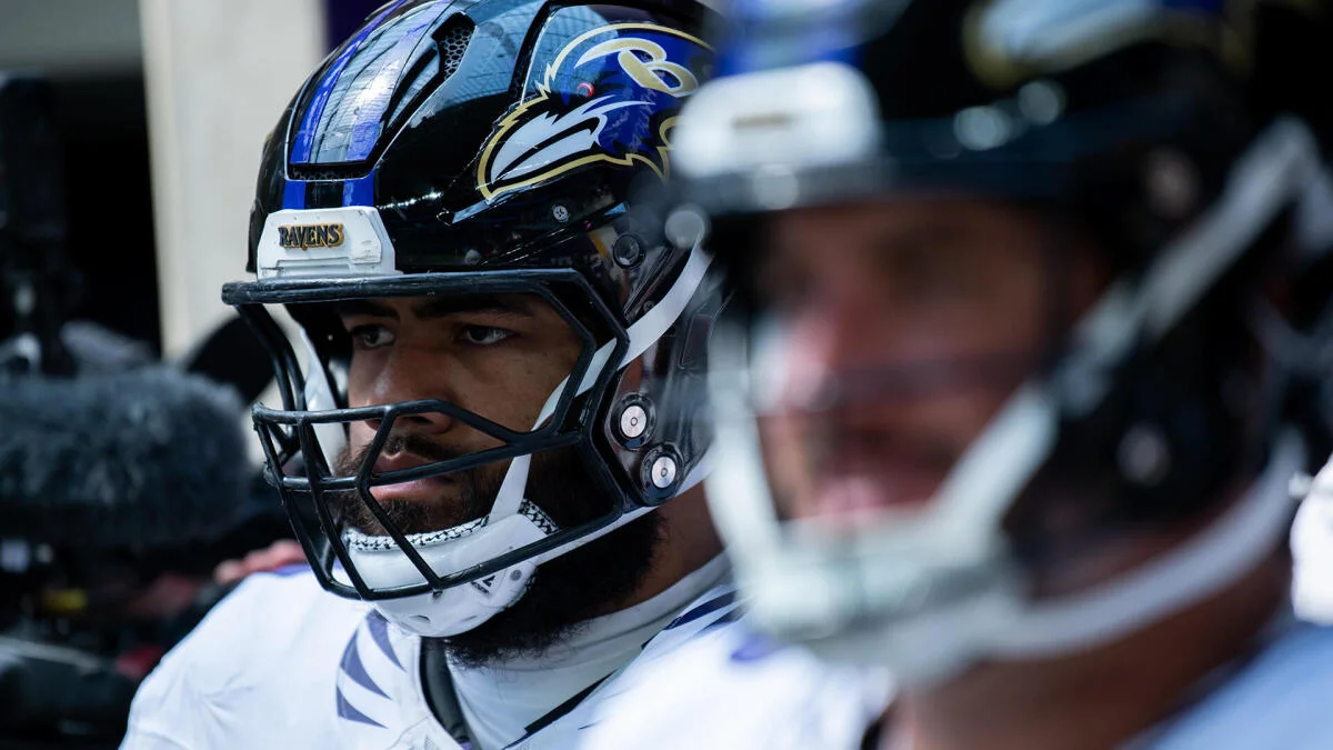 A Ravens player in a helmet sits on the sideline beside a blurred teammate.