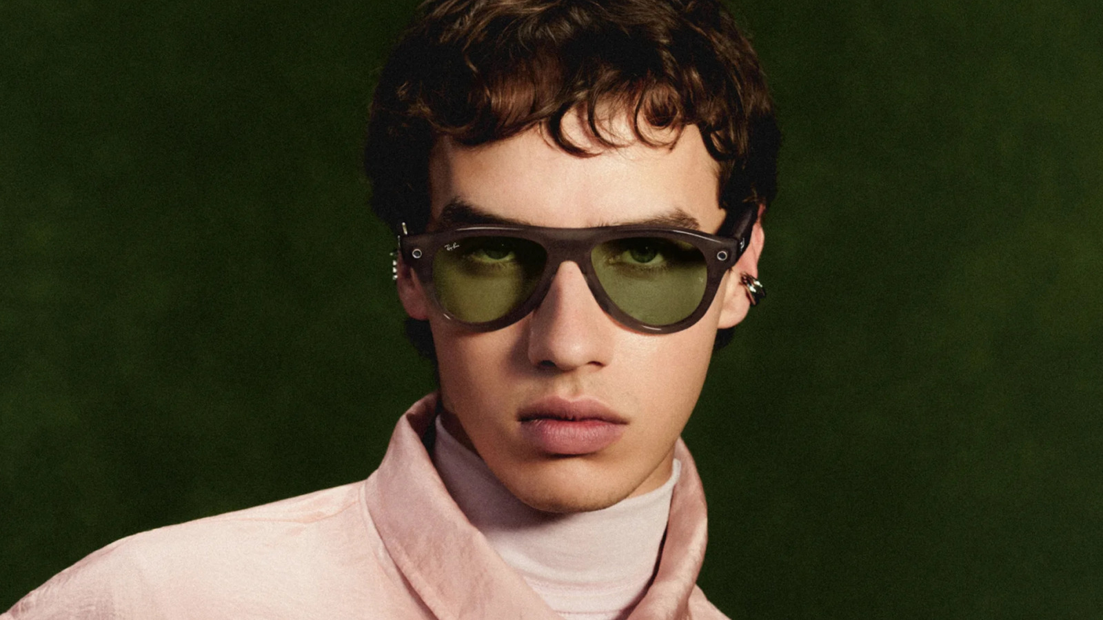 Close-up portrait of a person wearing dark smart glasses, a pale pink scarf, and a light jacket against a green background.
