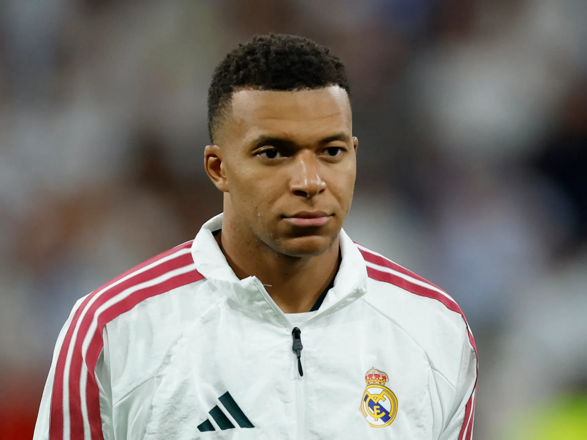 Footballer in a white Real Madrid jacket looking toward the camera in a blurred stadium setting