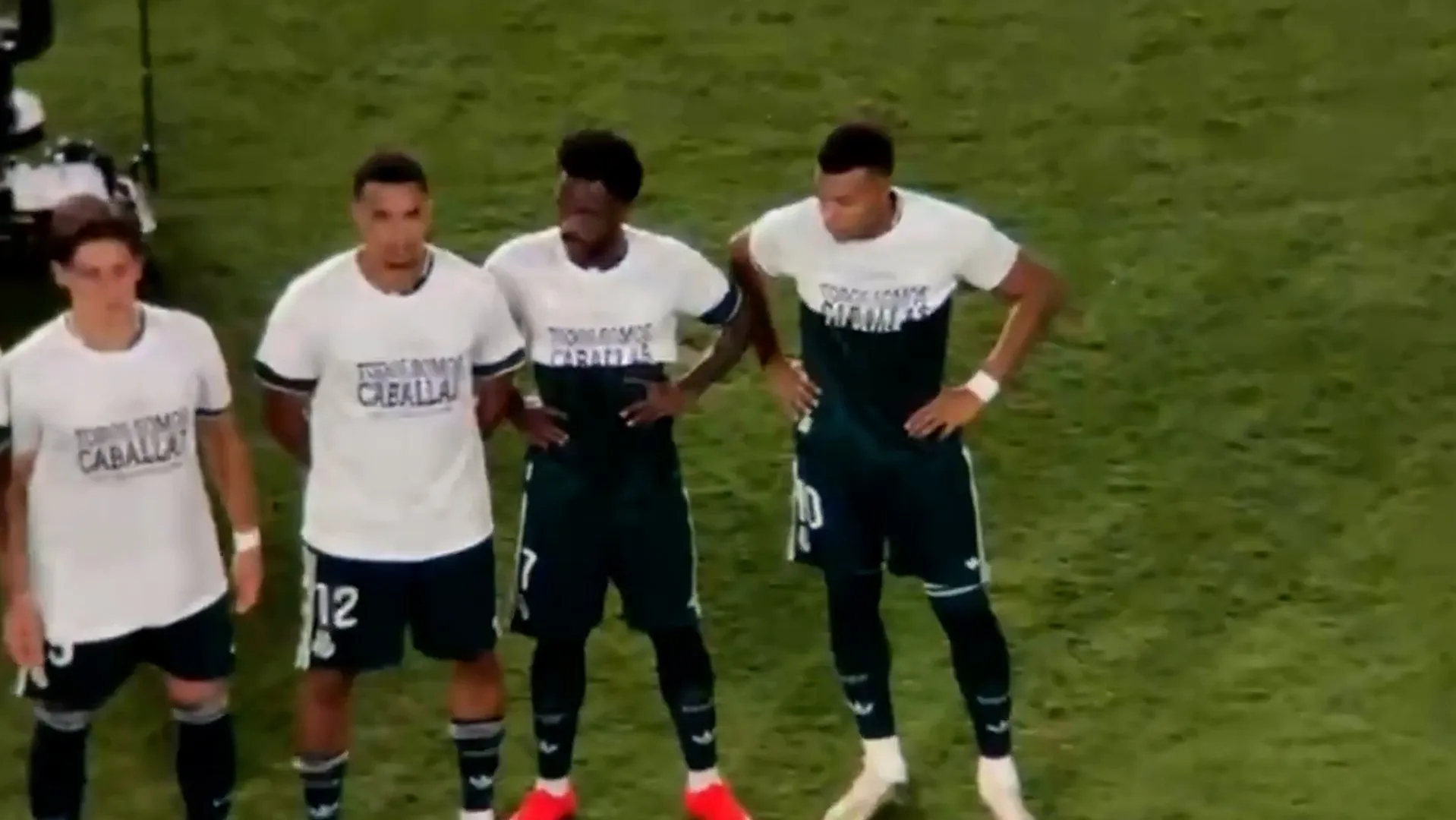 Football players stand on a grass pitch wearing white shirts over dark uniforms.