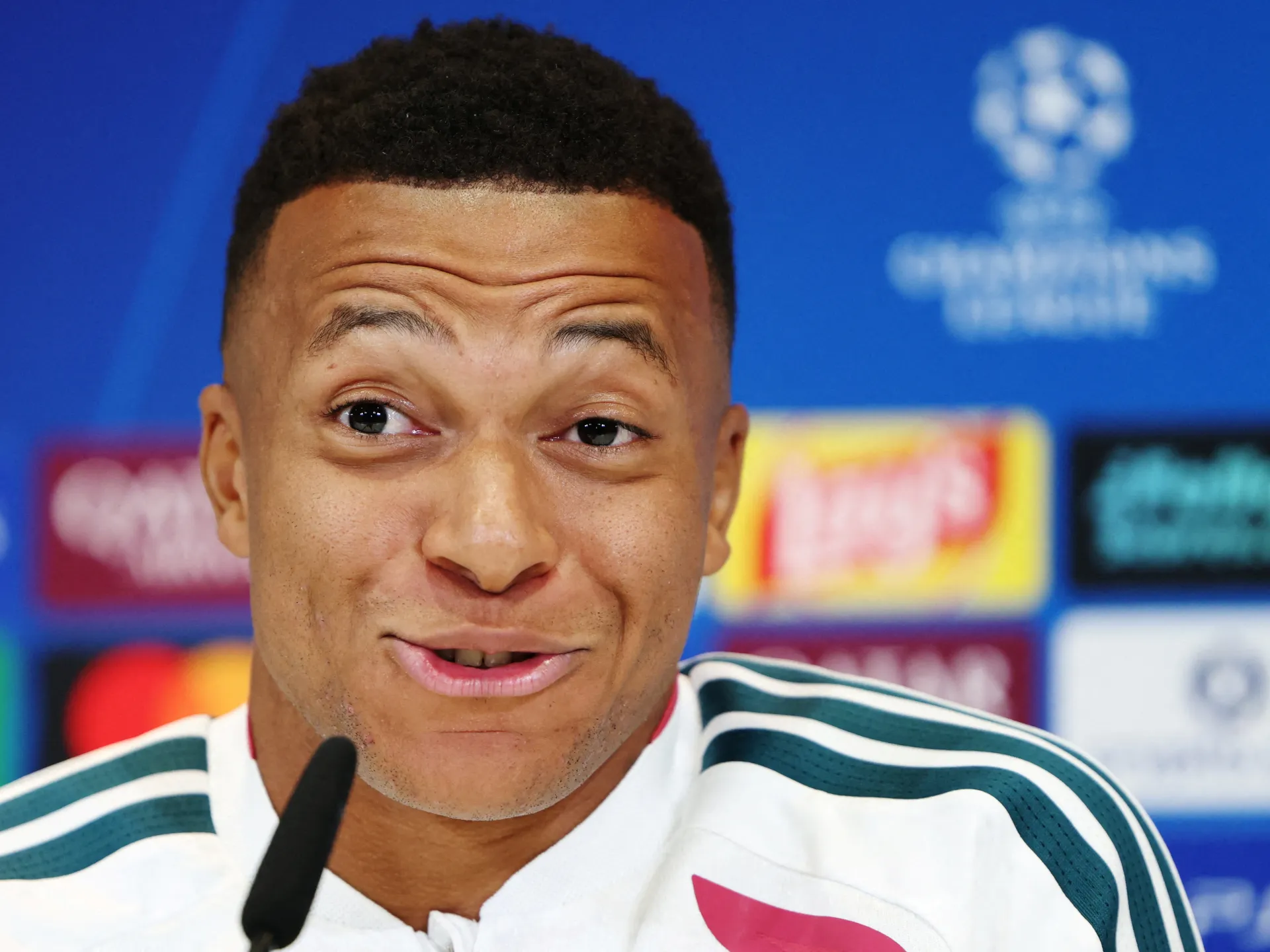 Footballer speaking into a microphone at a press conference, wearing a white and green-trimmed jacket.