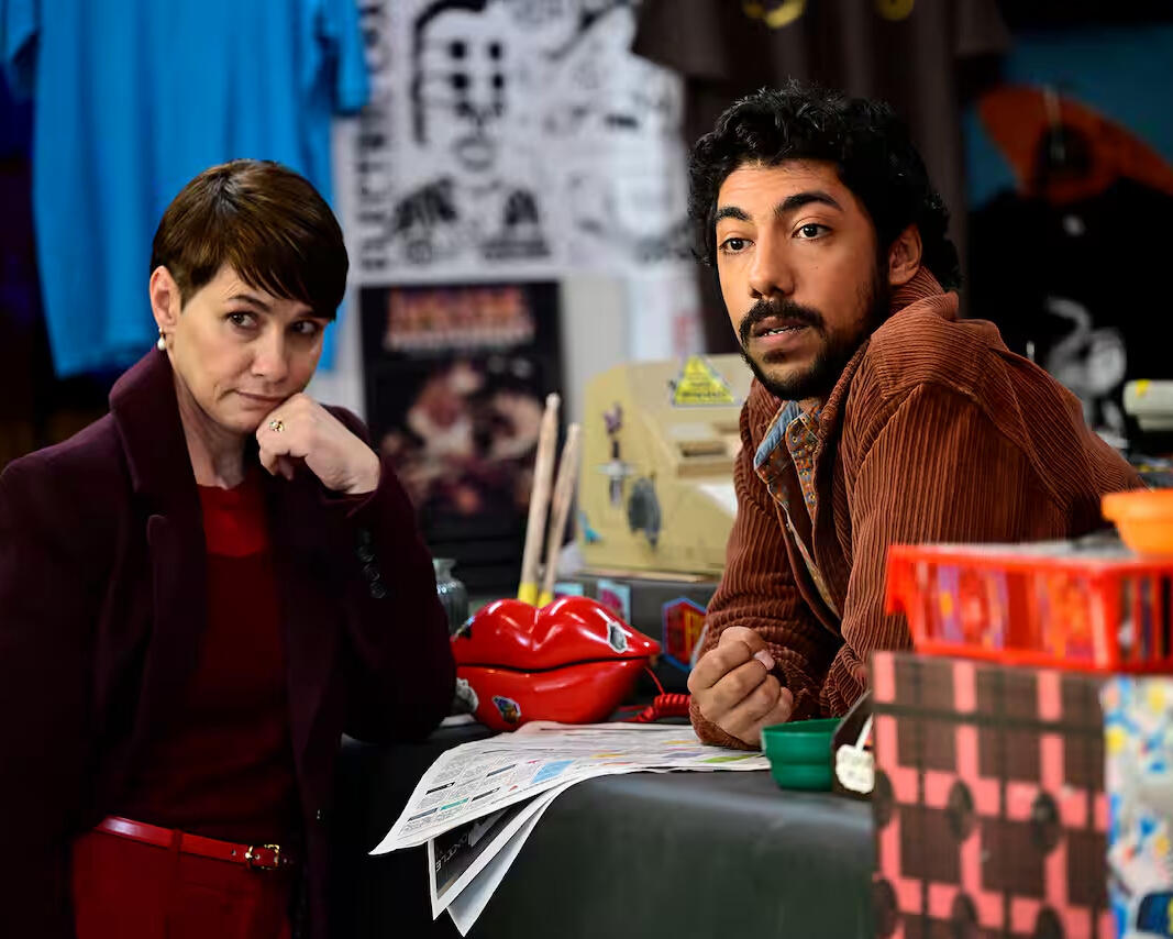 Two people sit at a cluttered table surrounded by posters, props, newspapers and a red lips telephone.