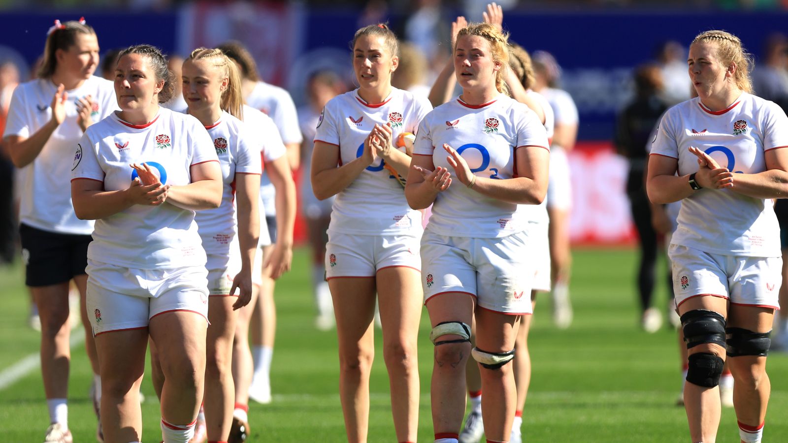 England women’s rugby players in white jerseys walk across the pitch applauding.