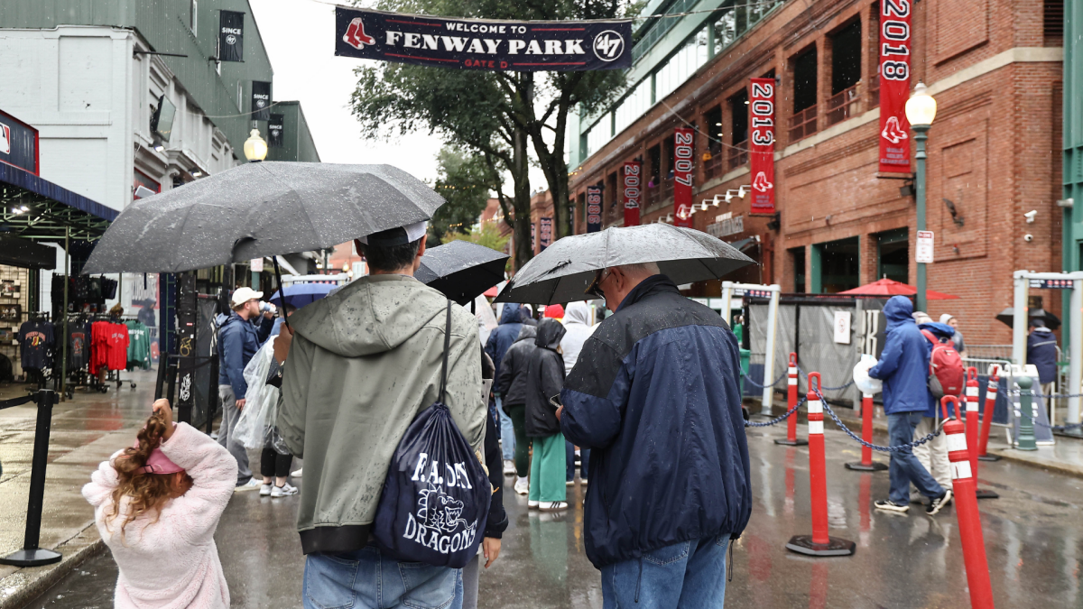 Fans carrying umbrellas walk along a rain-soaked stadium entrance beneath a Fenway Park banner.