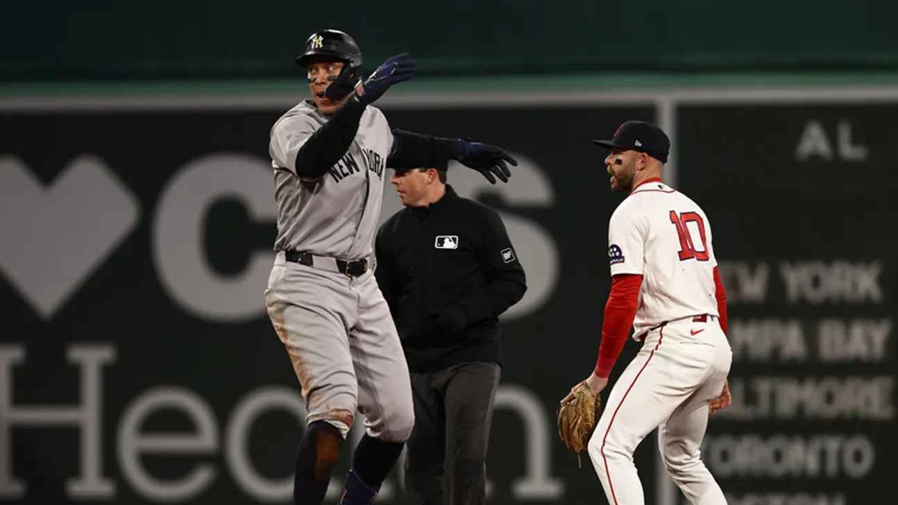 Yankees player gestures beside an umpire as a Red Sox player watches on the field.