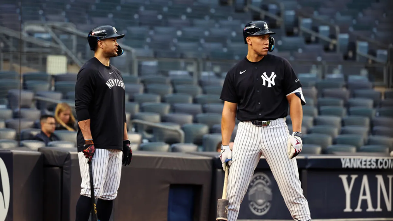 Two baseball players in Yankees uniforms stand near the dugout, with bats and empty seats behind them.