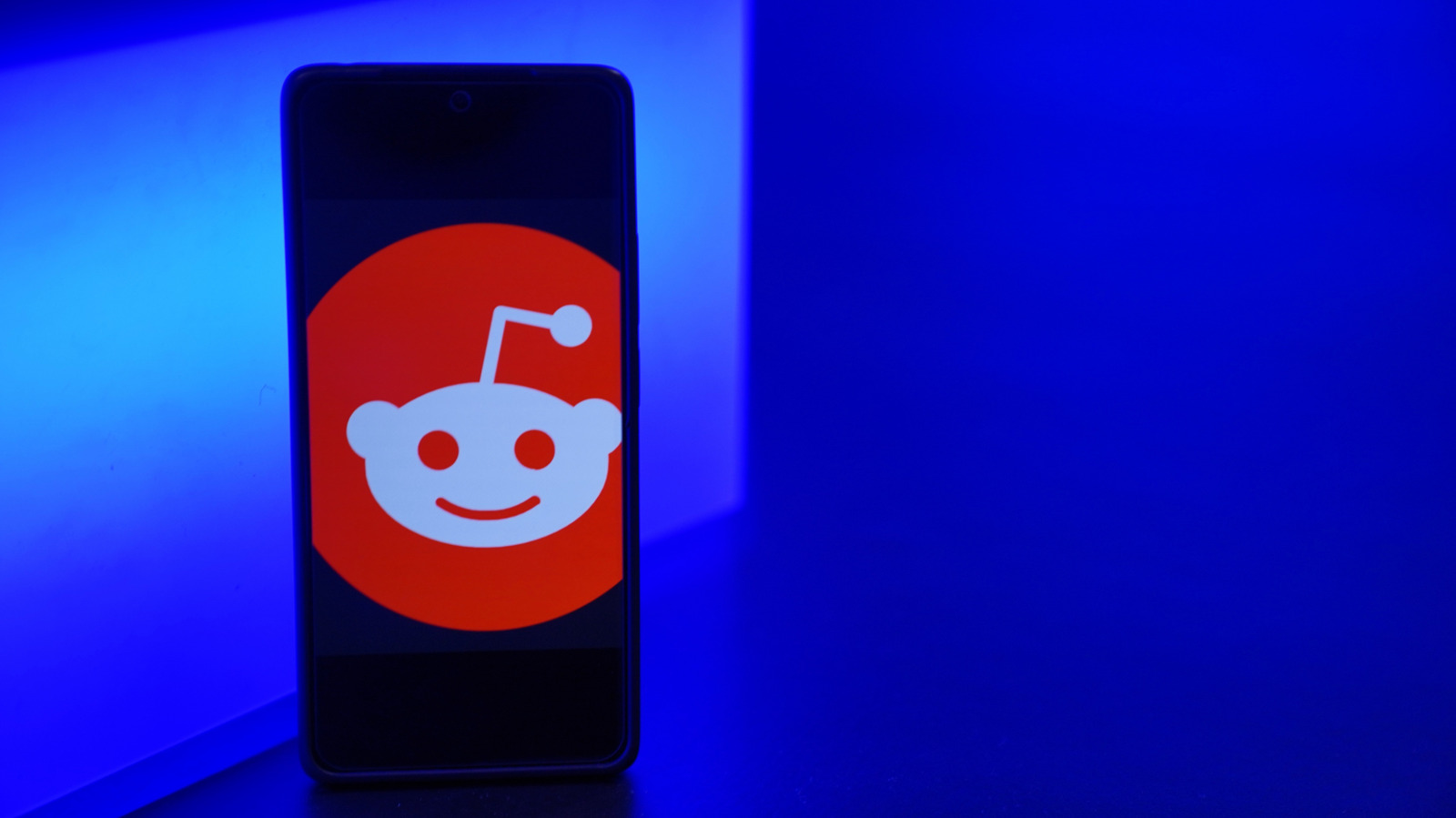 A smartphone displaying the Reddit logo stands against a brightly lit blue background.