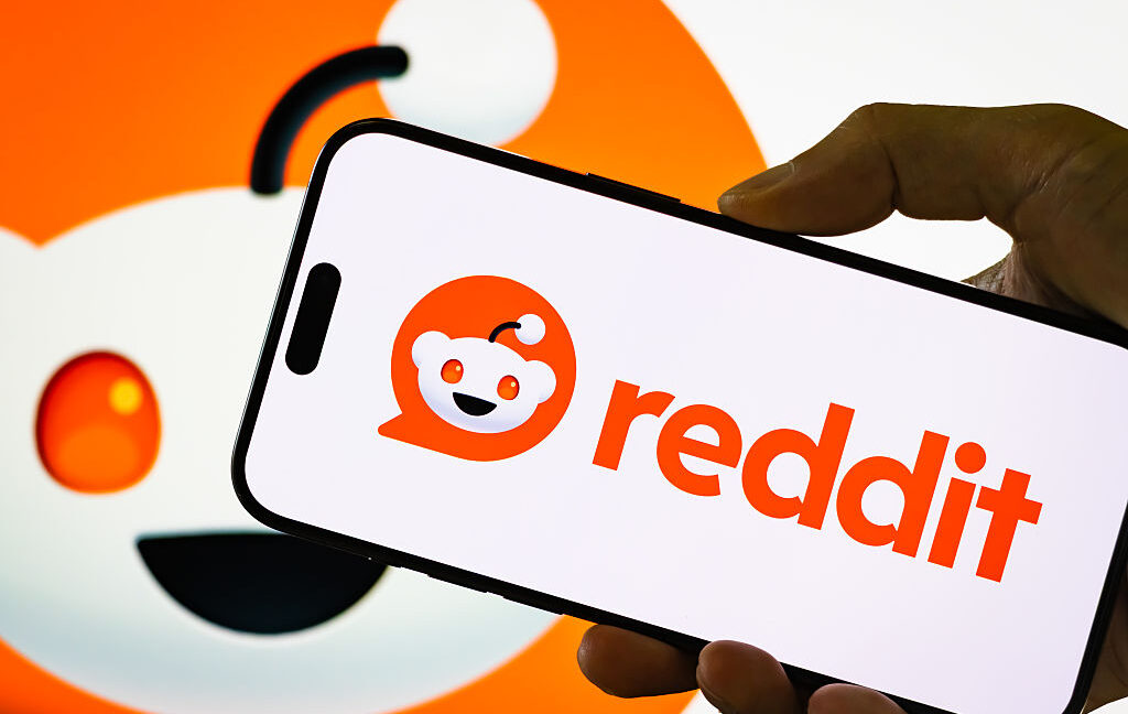 A hand holds a smartphone displaying Reddit’s logo and wordmark against a large Reddit logo backdrop.