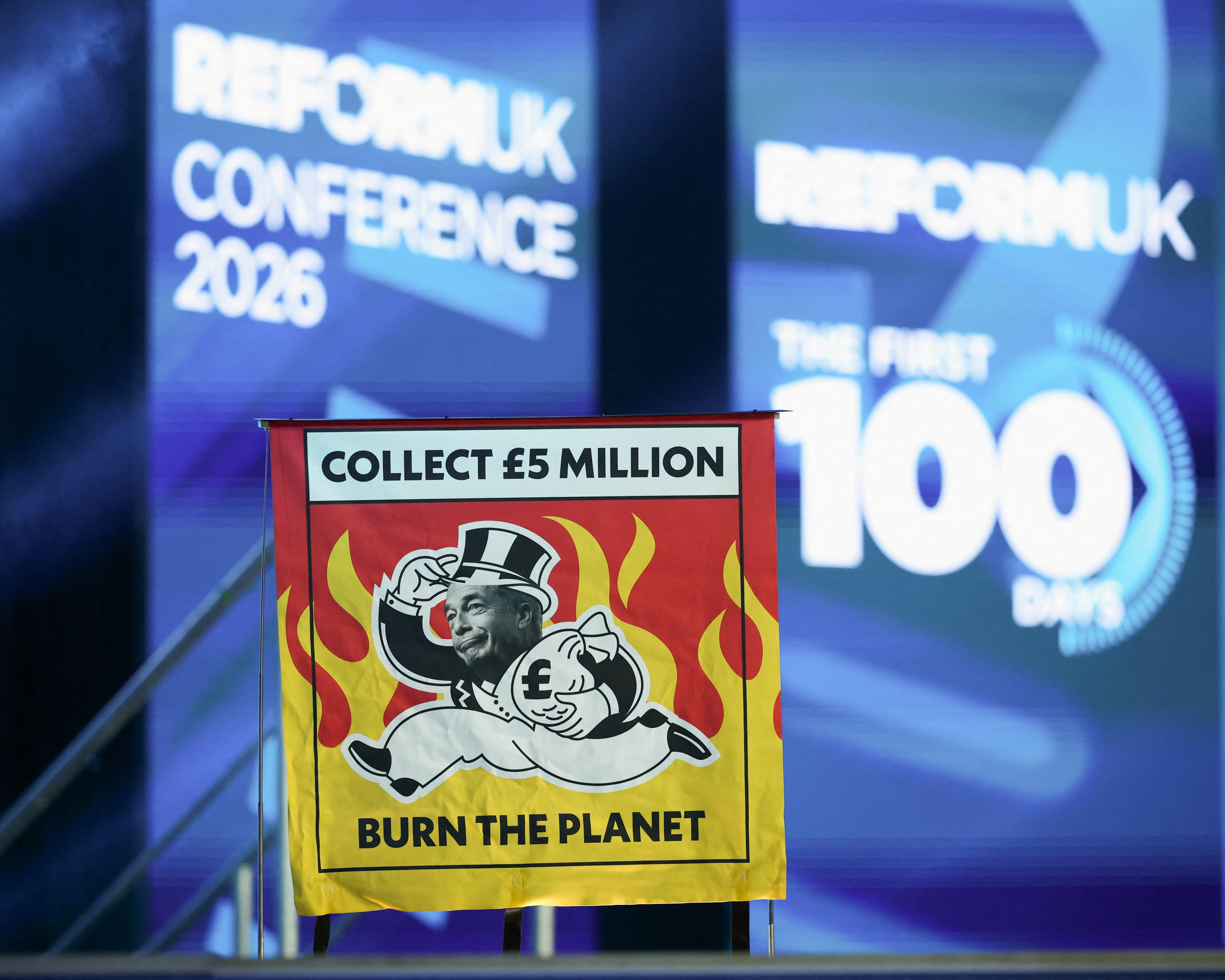 A placard reading “Collect £5 million” and “Burn the planet” at a blue-lit conference.