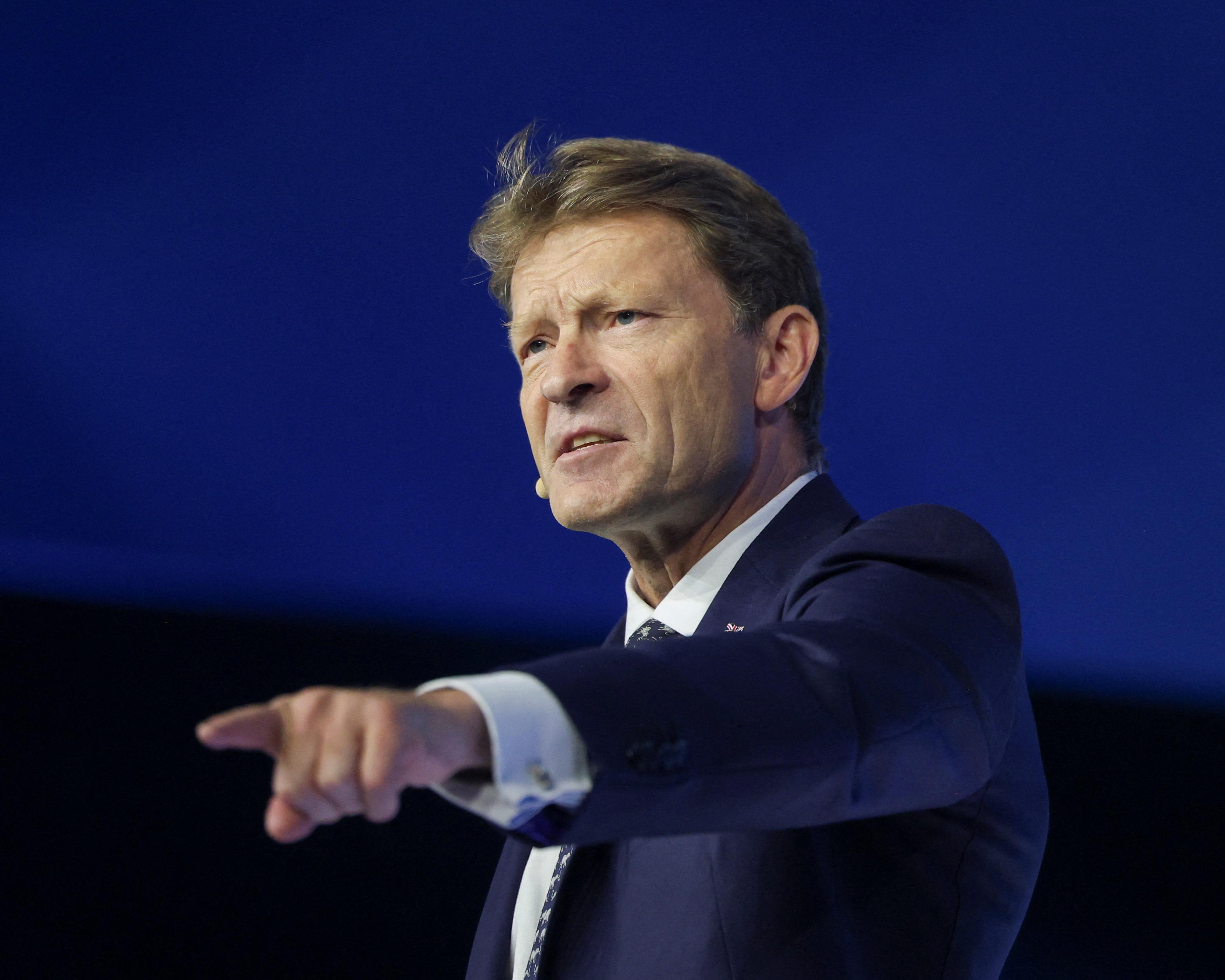 Man in a suit and tie points forward while speaking against a dark blue backdrop