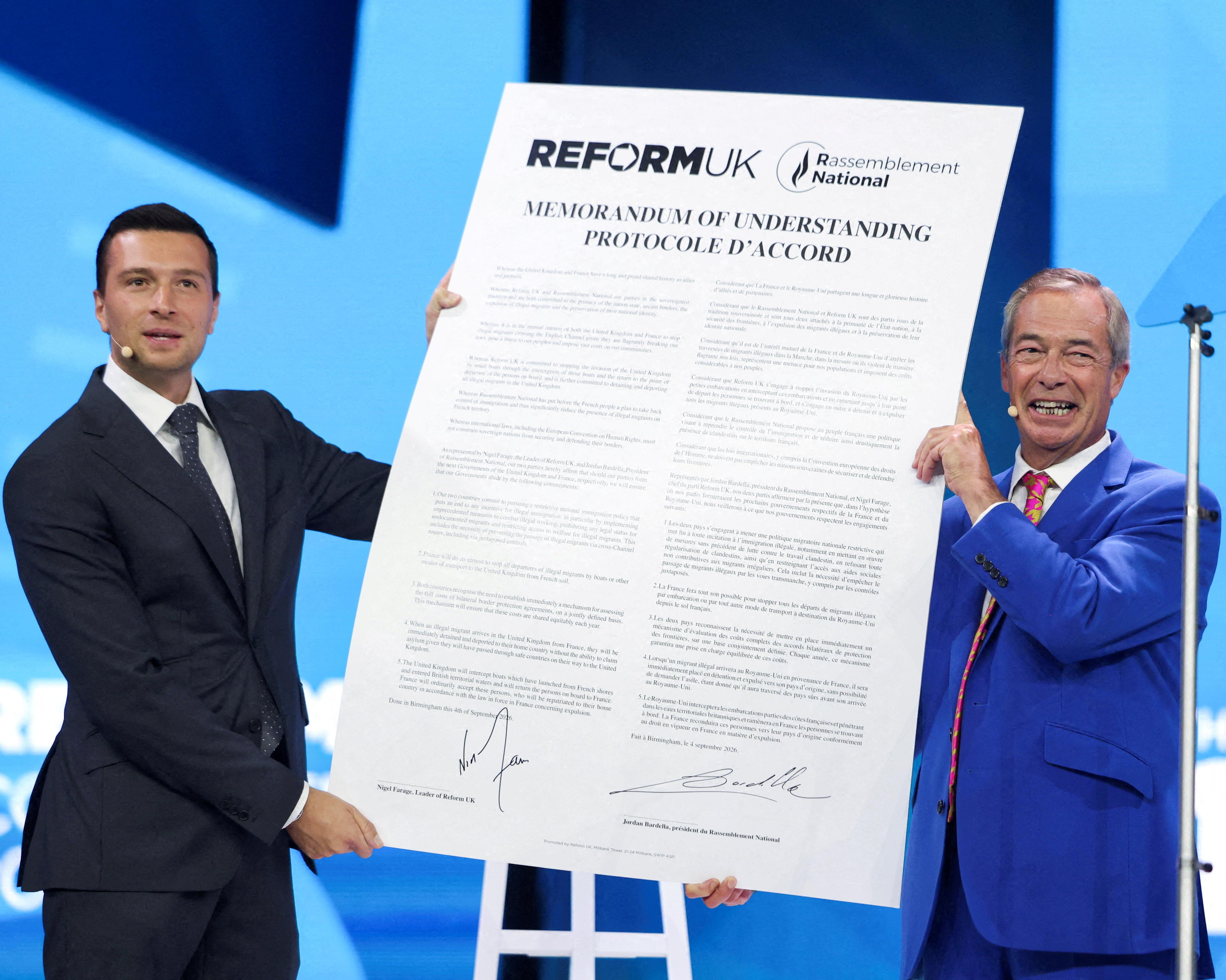 Two men hold a signed bilingual Reform UK and Rassemblement National memorandum onstage.