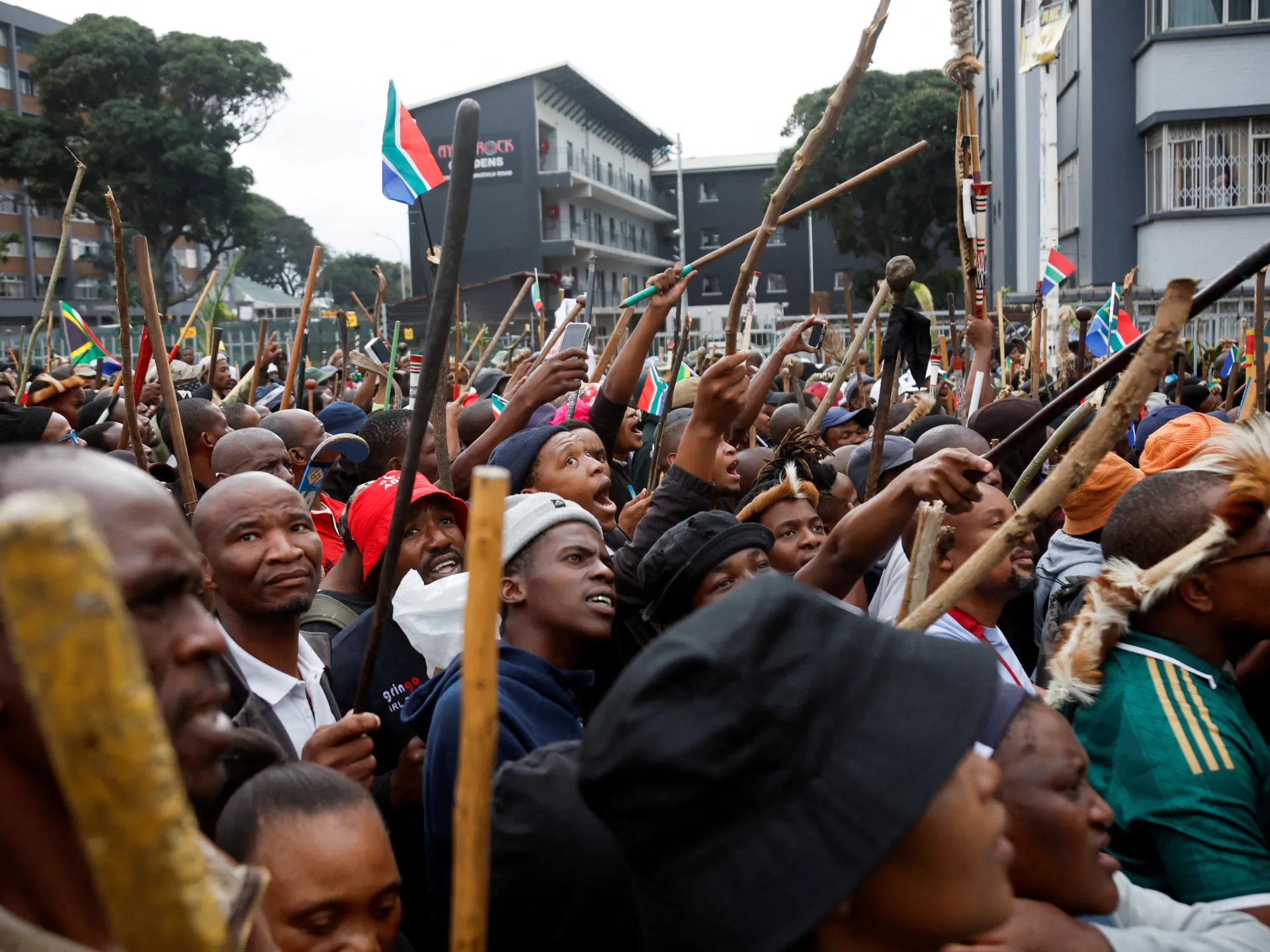 Crowd of people holding sticks and South African flags outside urban buildings.