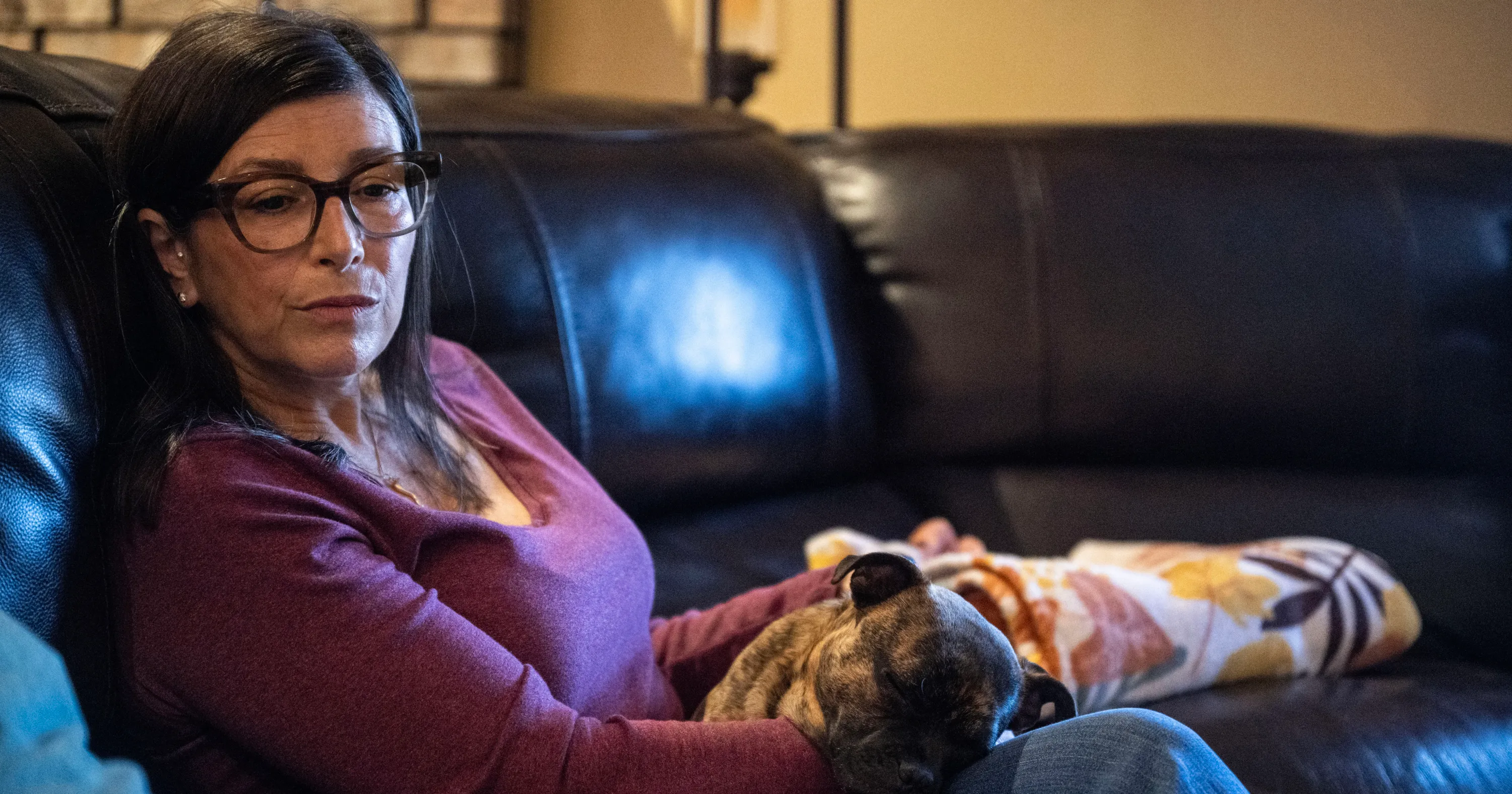 Woman wearing glasses sits on a couch holding a sleeping dog in her lap.