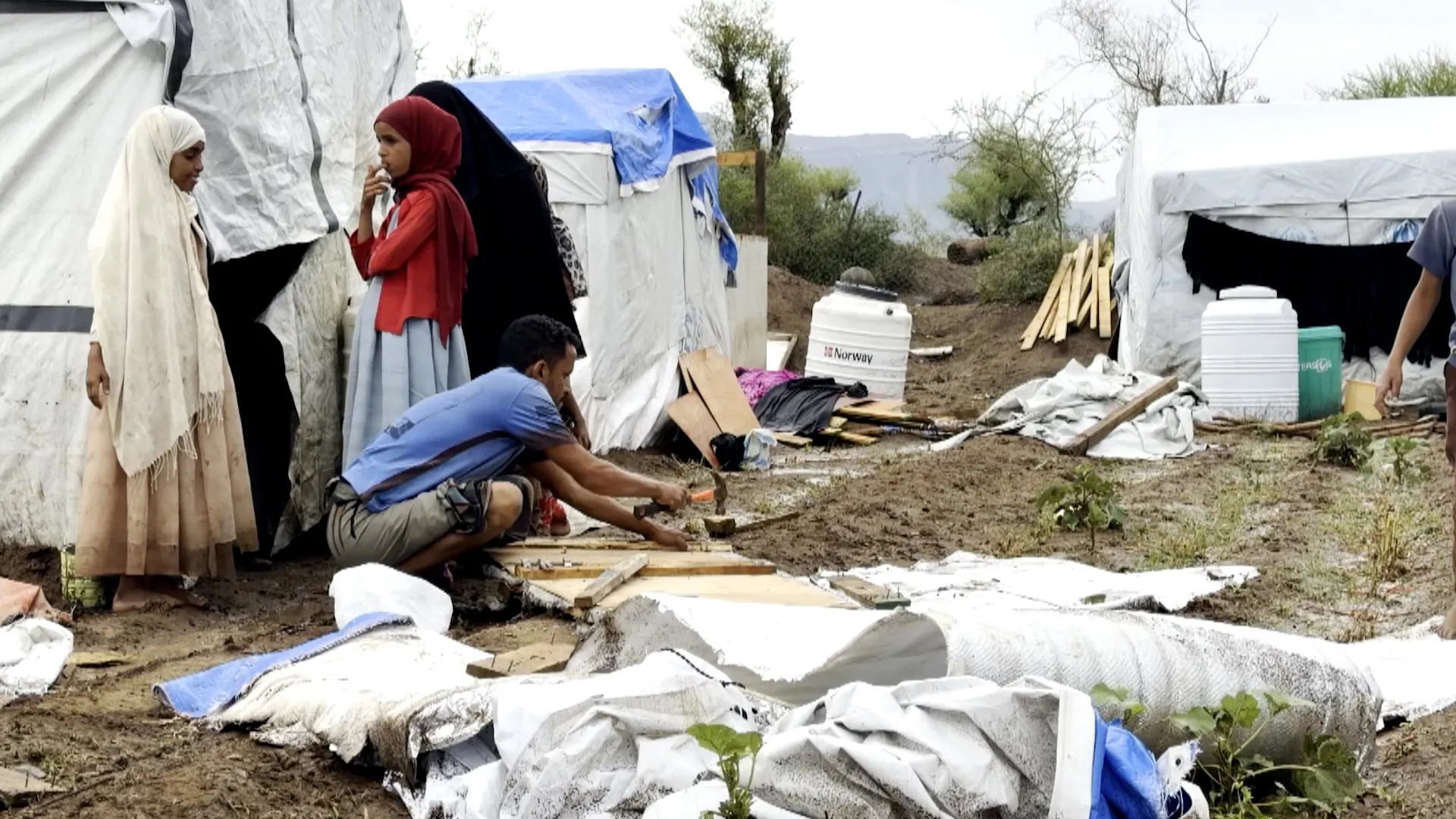 People repair makeshift tents beside water containers and scattered building materials.
