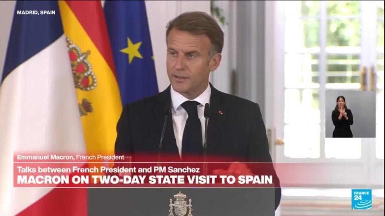 A suited man speaks at a lectern before French, Spanish, and EU flags, with a sign-language interpreter inset.