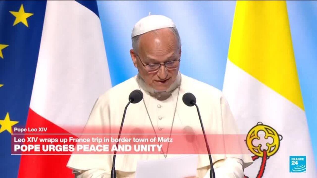 A white-robed cleric reads at a lectern between French and Vatican flags, with microphones in front.