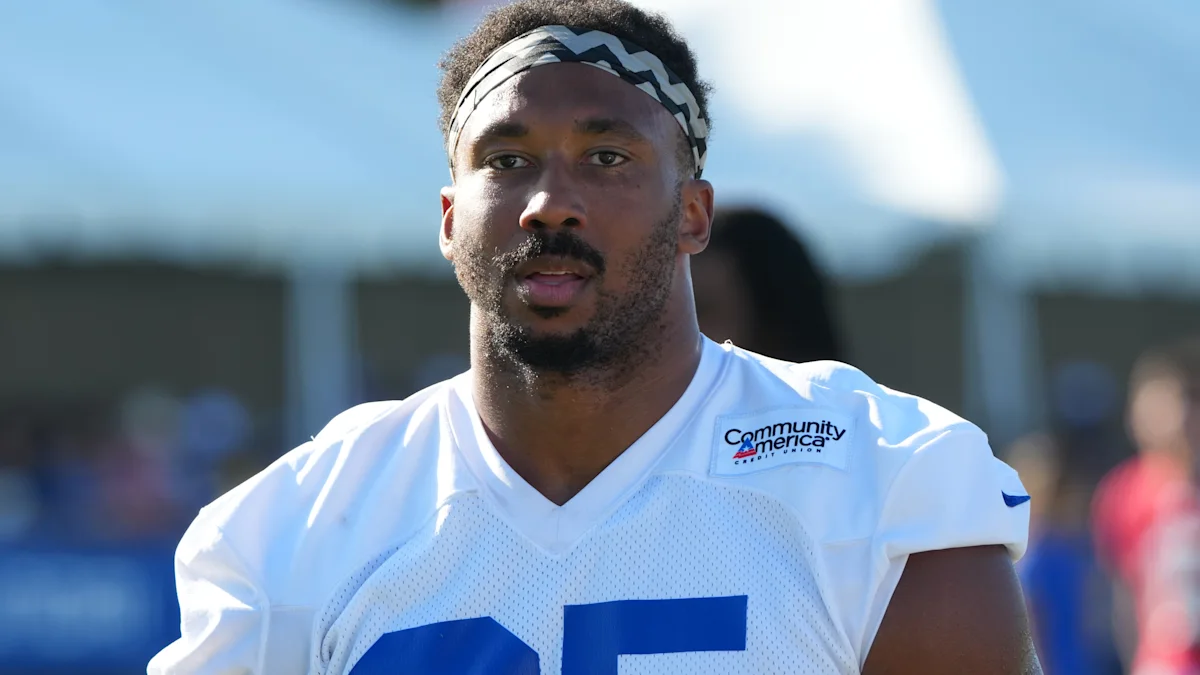 Football player in a white practice jersey and patterned headband stands on an outdoor field.
