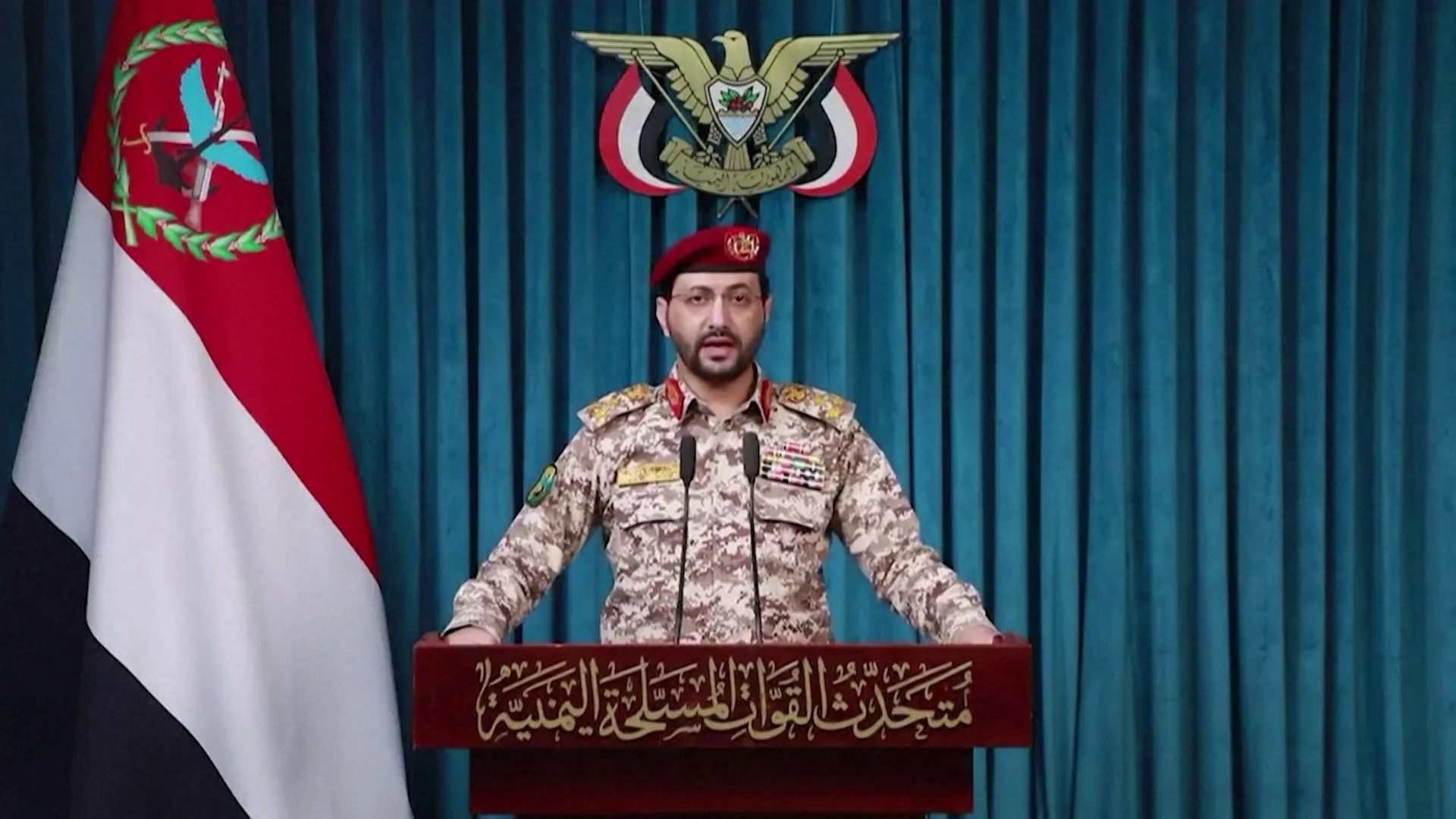 Uniformed military spokesman speaks at a podium before a Yemeni flag and national emblem.