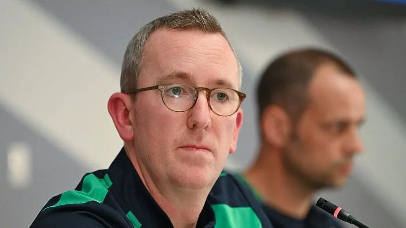 Man wearing glasses and a green-and-black top sits at a press conference beside a microphone.