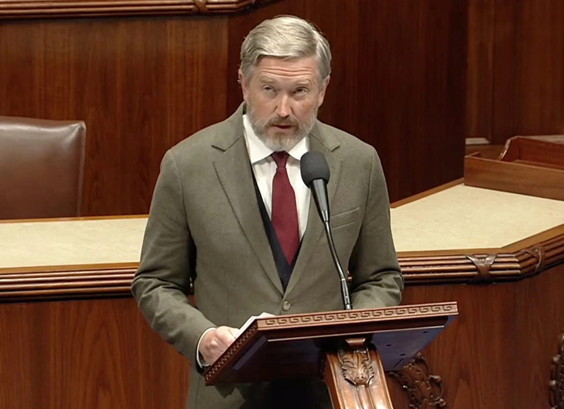 Gray-haired man in suit speaking at a lectern with a microphone in a wood-paneled chamber