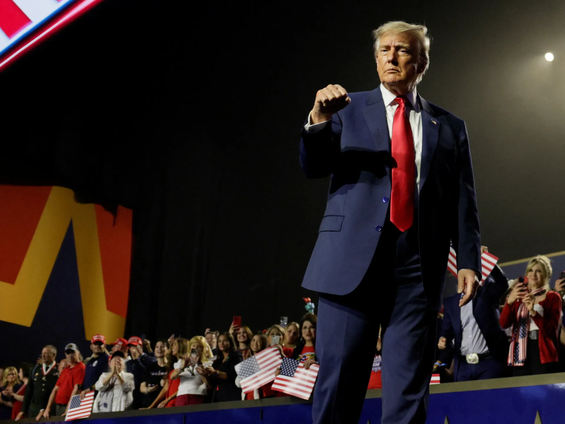 Donald Trump raises a fist onstage before a crowd holding American flags.