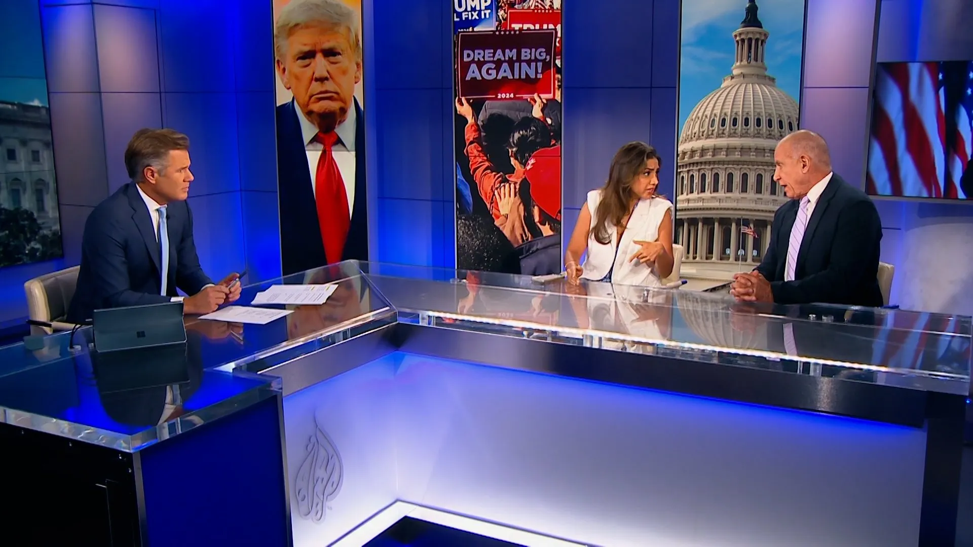 Television news panel with three commentators seated at a glass desk before political campaign images.