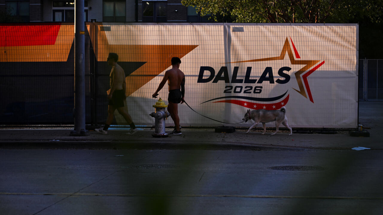 Two pedestrians walk a leashed dog past a Dallas 2026 event mural at dusk.