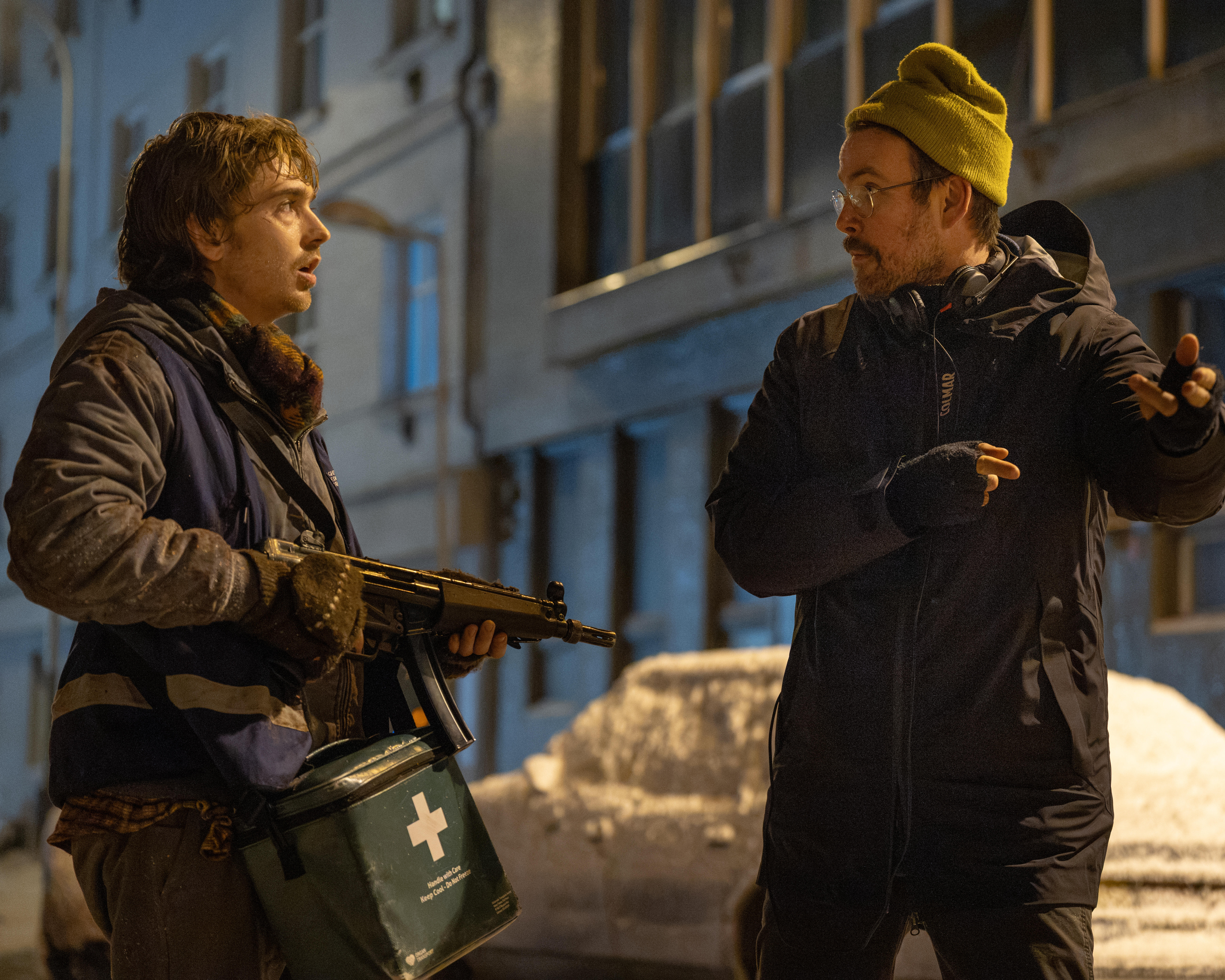 Two men discuss a scene outdoors at night; one holds a rifle and medical supply container.