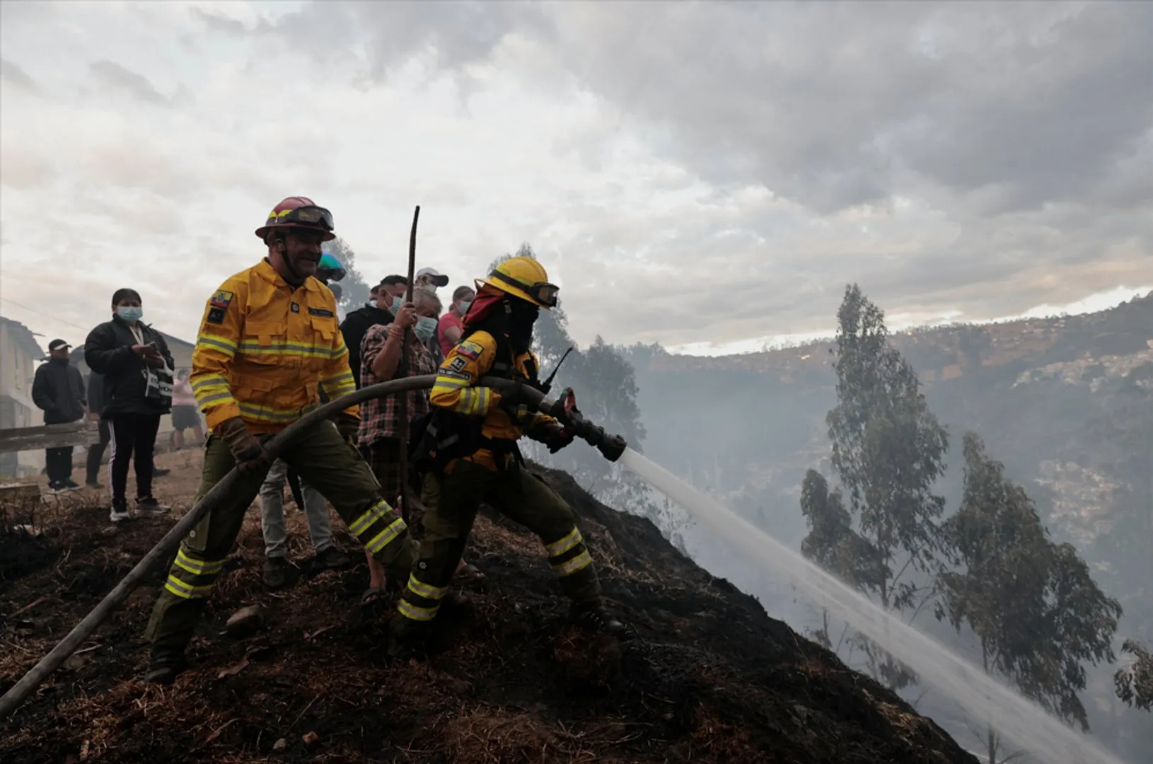 Firefighters and residents battle a smoky hillside blaze with a hose near homes.