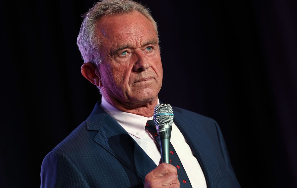 Older man in a dark suit holding a microphone against a black background