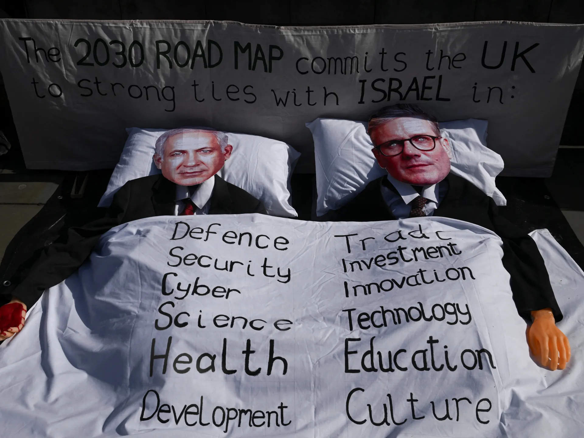 Staged display shows two suited figures lying in beds beneath handwritten UK-Israel relationship slogans.