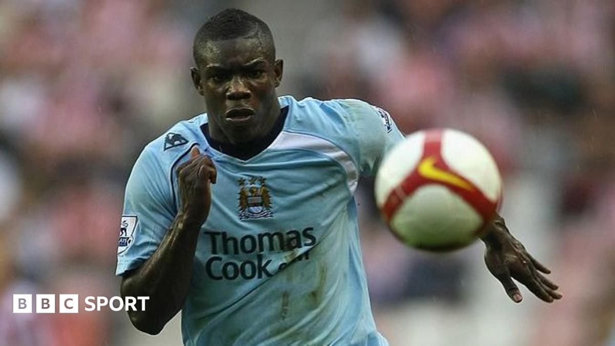 A Manchester City player in a light blue shirt runs toward a football in the foreground.
