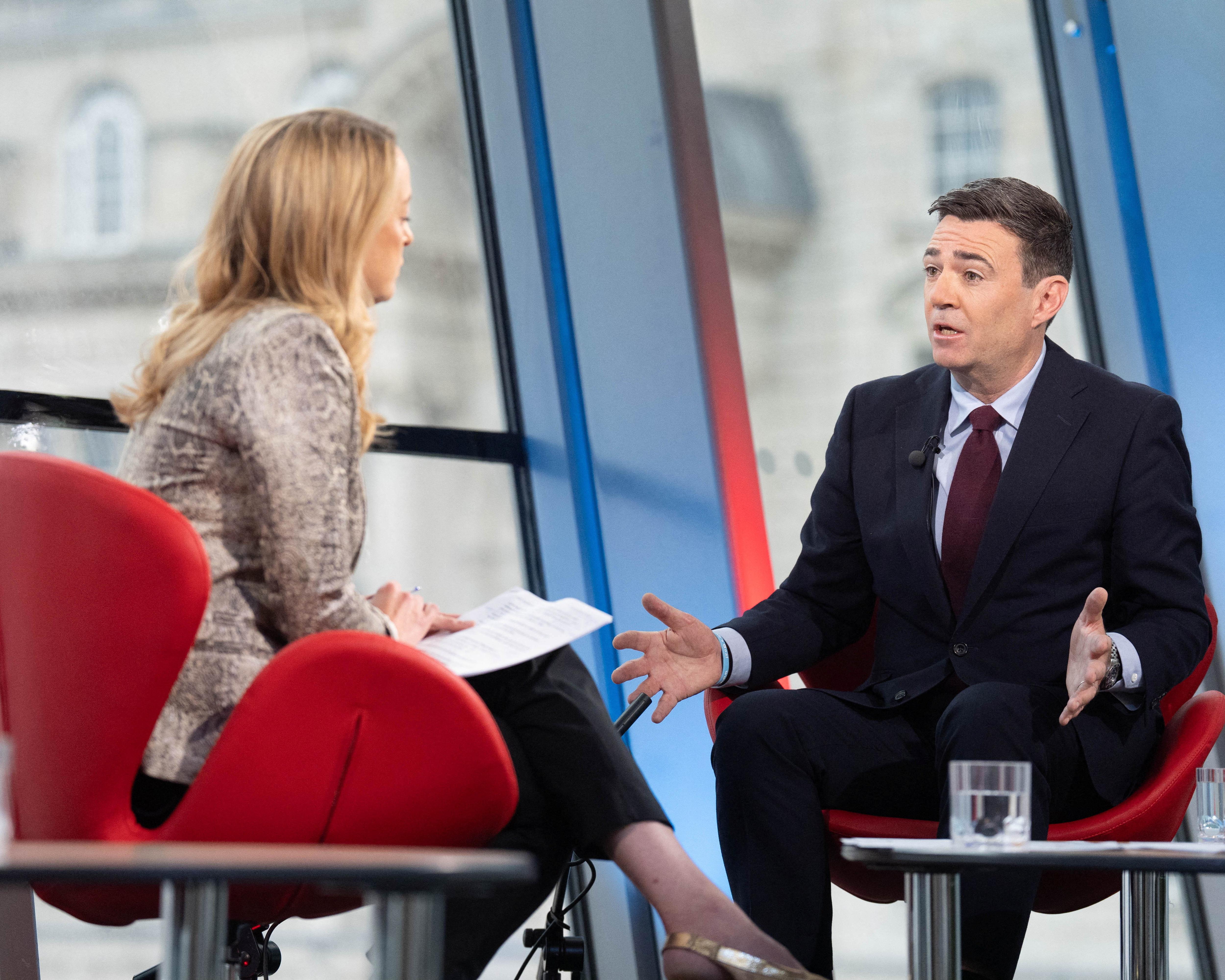 A suited man gestures while speaking to a woman holding papers, seated in red chairs on a studio set.