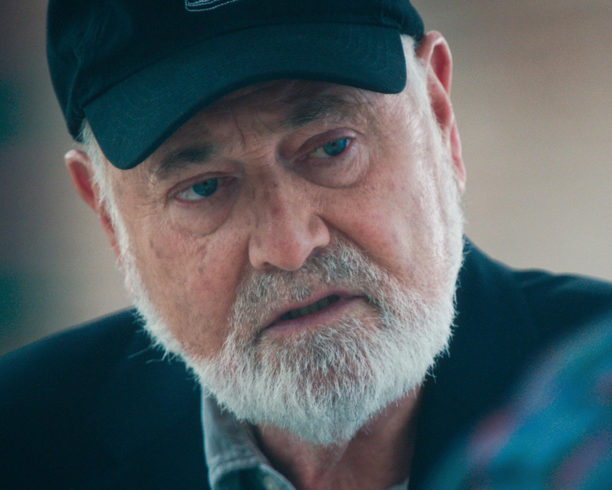 Close-up portrait of a bearded older man wearing a dark cap and jacket